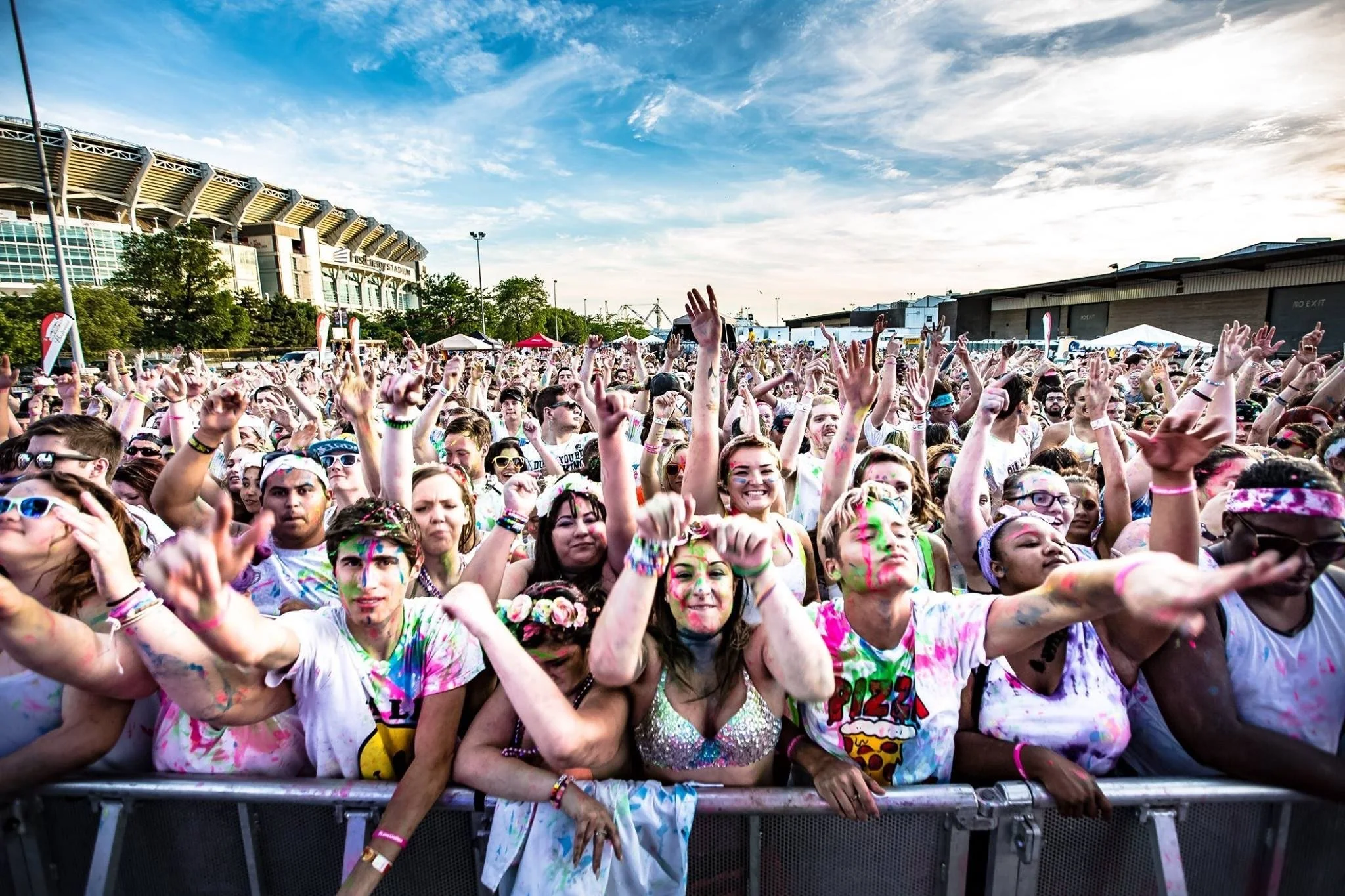 Crowd of people at a music festival or outdoor concert, covered in colorful paint and wearing casual attire, standing behind a barrier and enjoying the event under a bright sky.