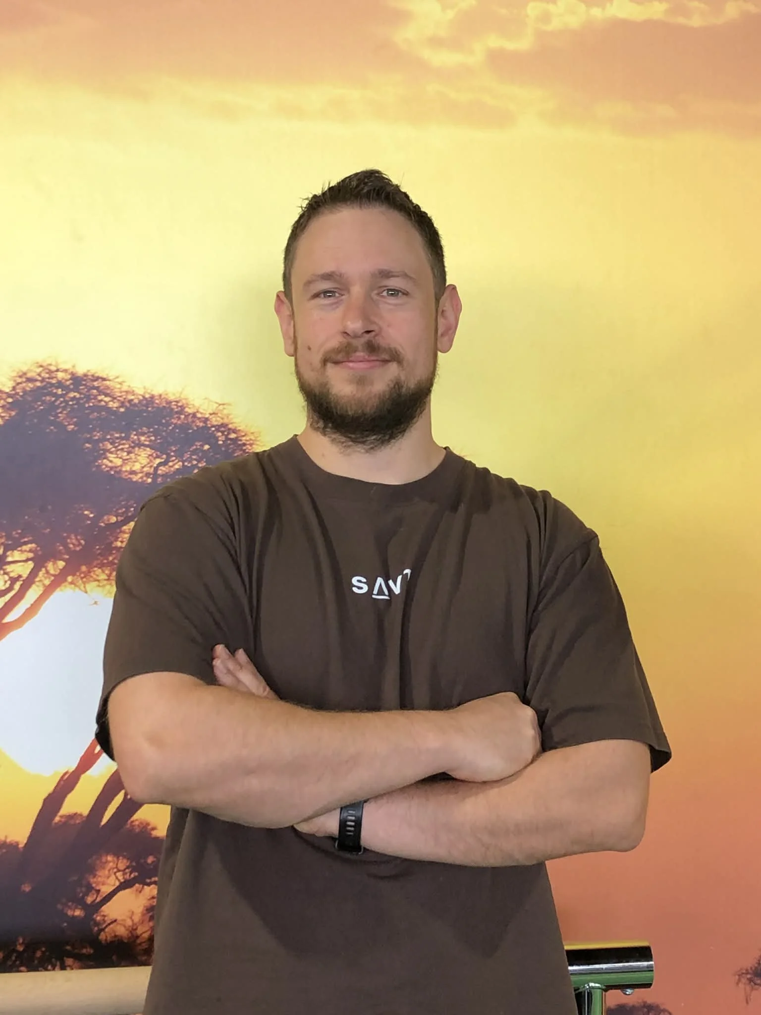 A man standing with arms crossed in front of a sunset African savannah landscape background.