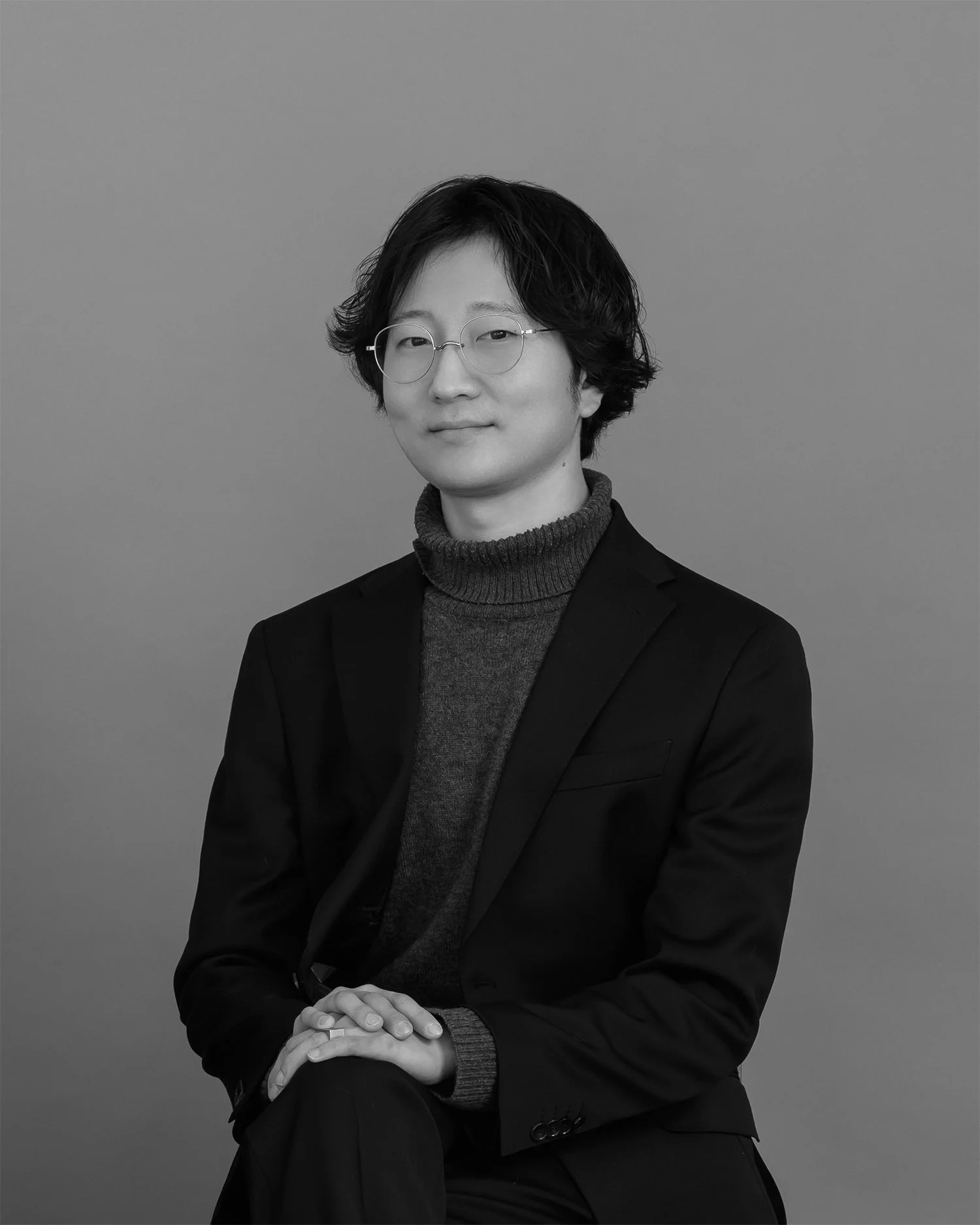 A black-and-white portrait of a person with medium-length dark hair, glasses, and wearing a turtleneck sweater and blazer, seated with hands resting on their lap against a plain background.