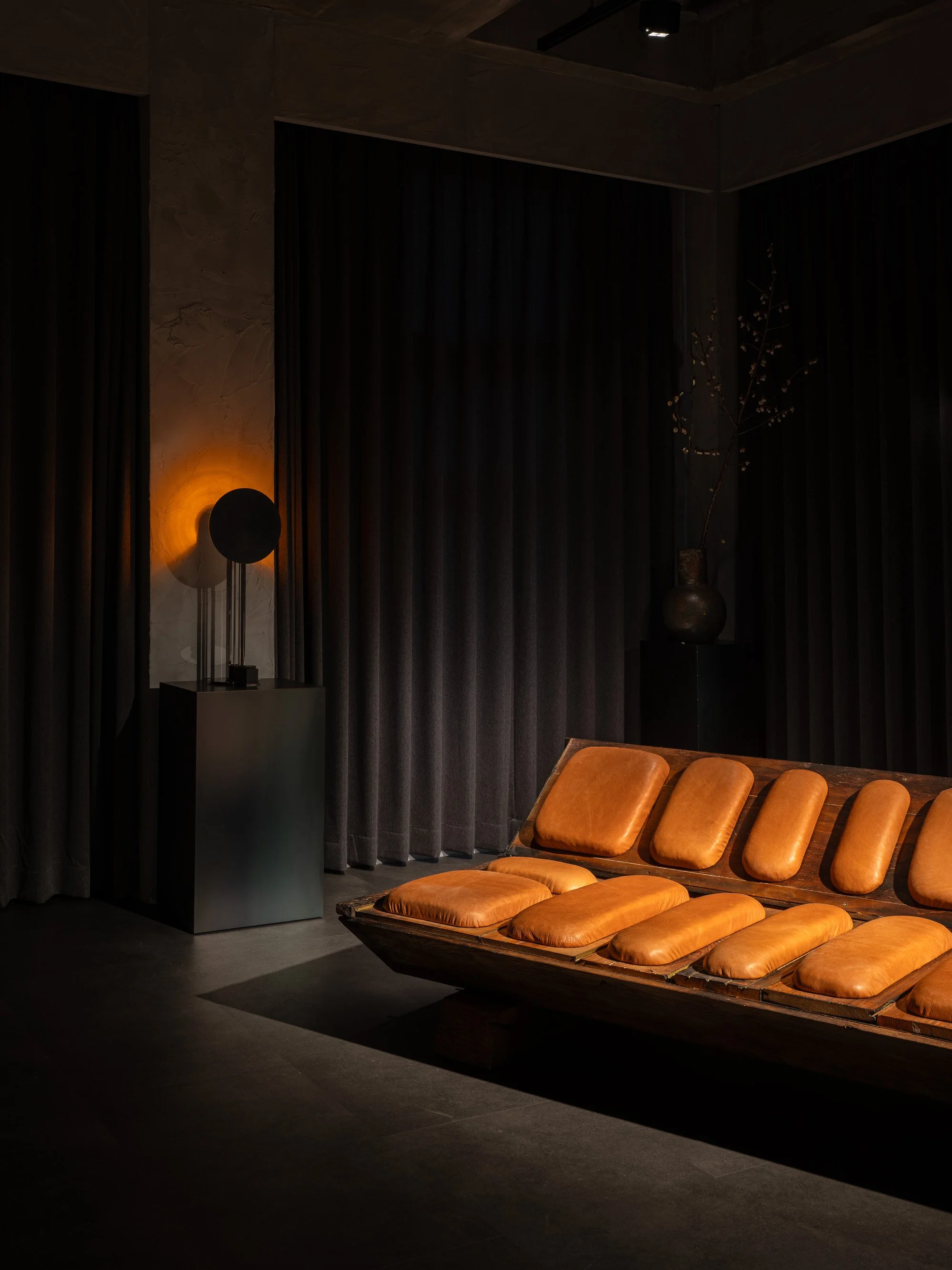 A dimly lit room with dark curtains, a modern sculpture lamp on a black pedestal, a tall ceramic vase with branches, and a wooden bench with brown leather cushions.