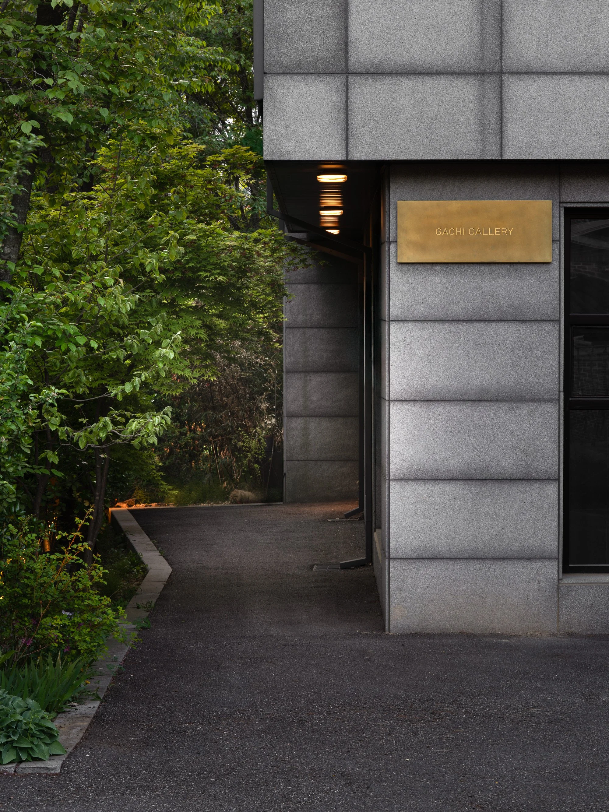 Entrance to Gachi Gallery, a building with gray concrete walls and a gold sign, surrounded by green trees and plants, with a dark asphalt pathway leading to the entrance.