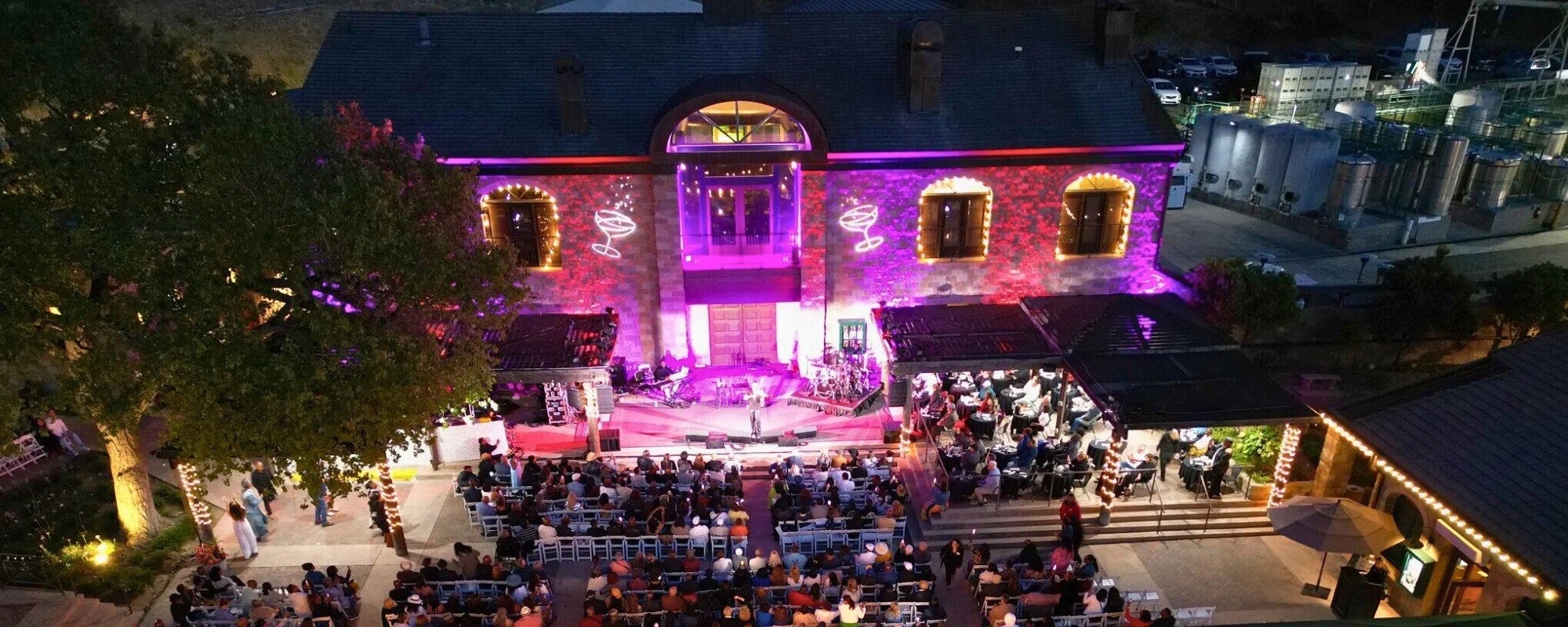 Thornton Winery Champagne Jazz Series nighttime outdoor concert with audience seated in front of a lit brick building decorated with purple and yellow lights, featuring neon signs of martini glasses on the wall.