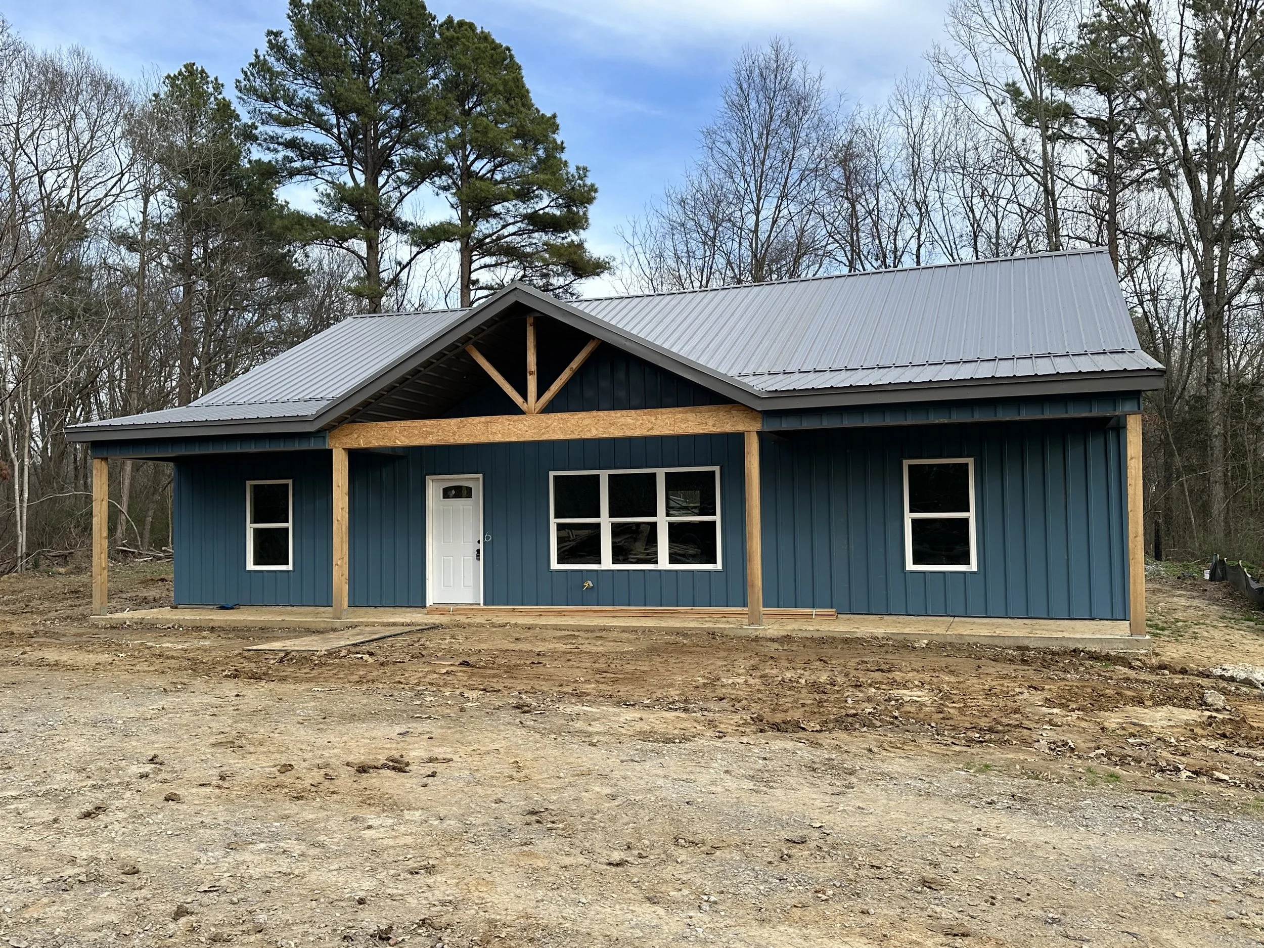Front porch of a house with a stone and siding exterior, wooden front door, and red support columns, surrounded by grass and trees.