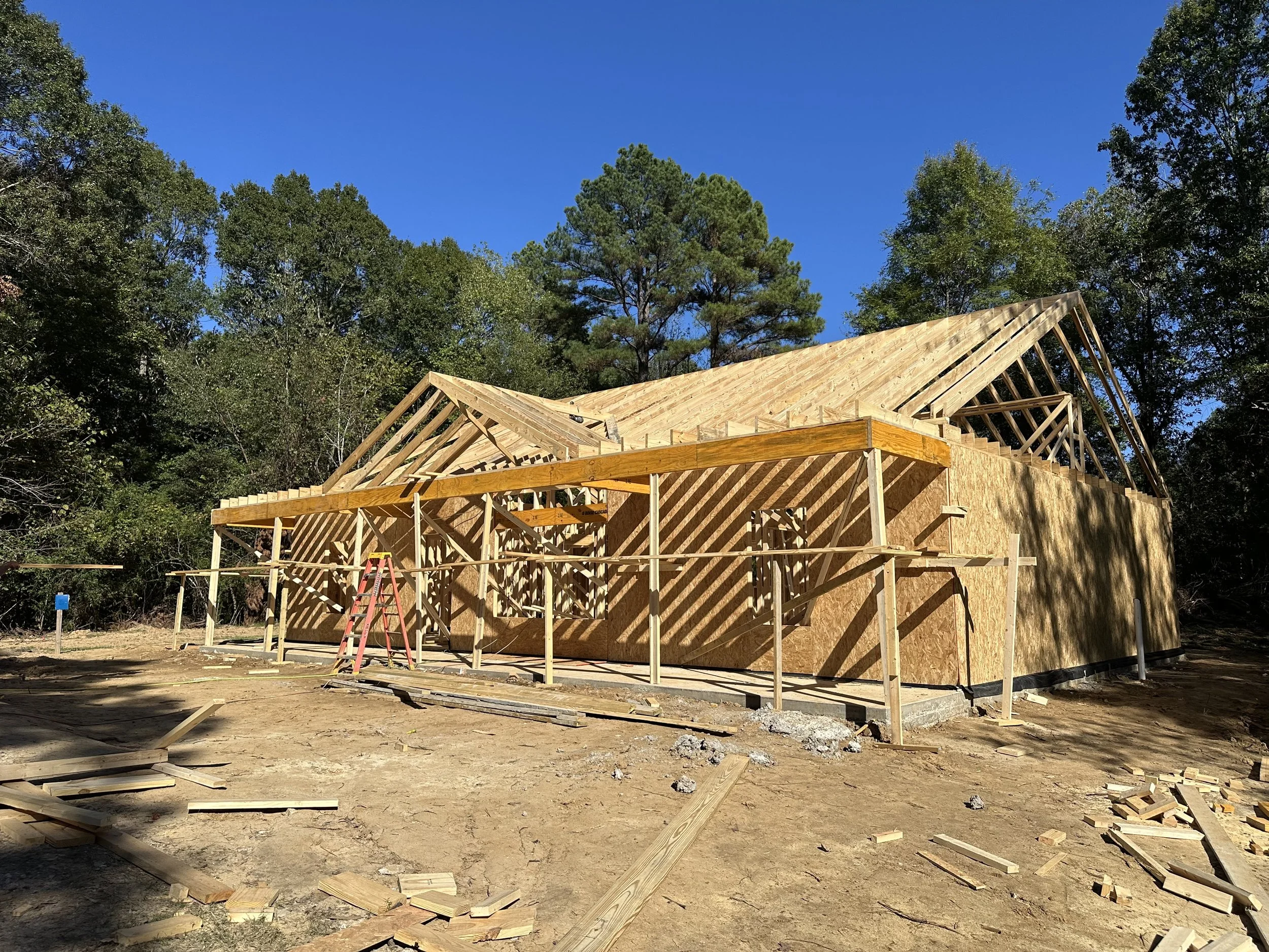 A house under construction with a metal roof, wrapped in building material, with a porch area and a large front window. There is a utility vehicle to the left and construction equipment in front.