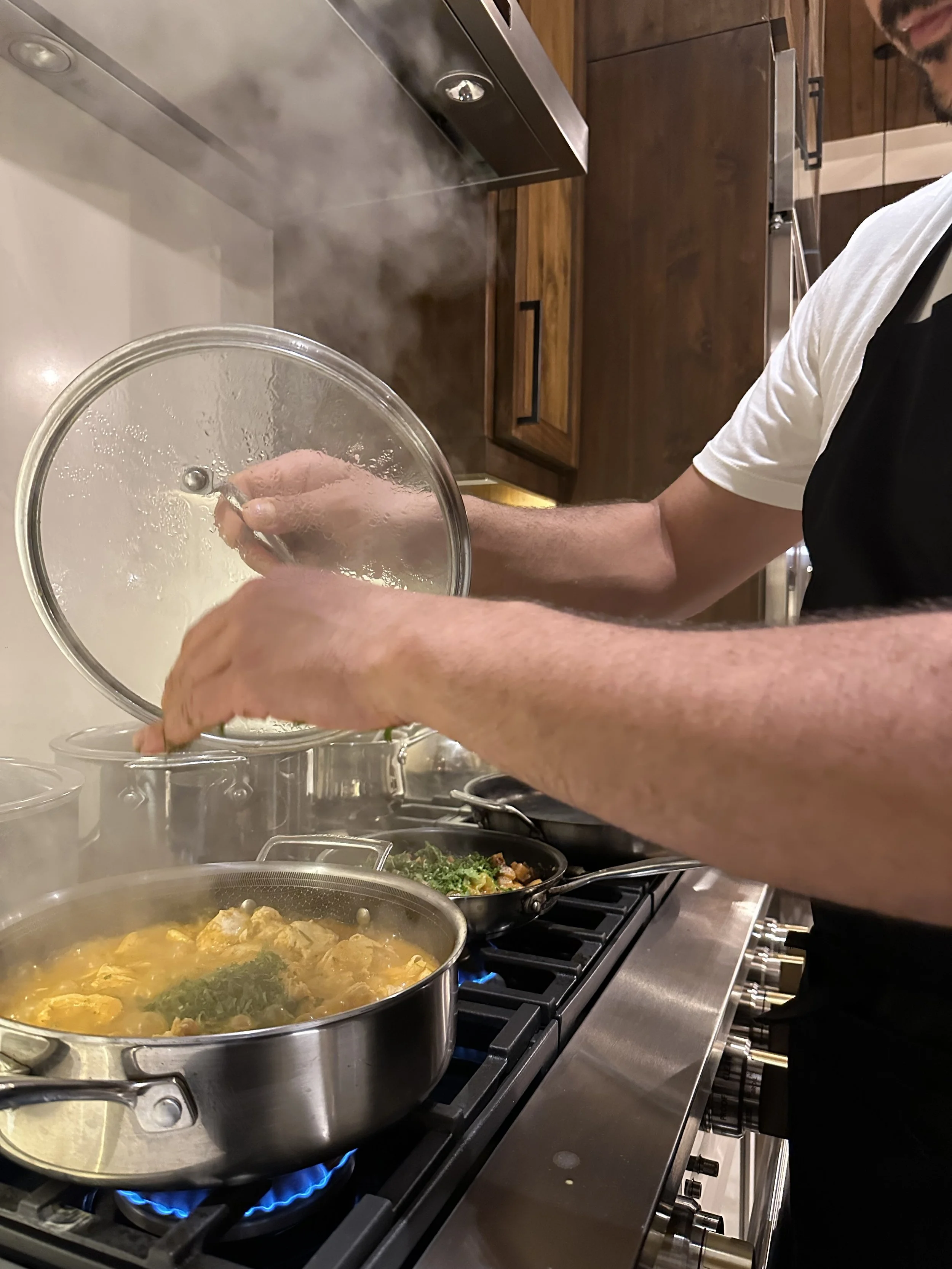 A person cooking on a stovetop, stirring a pot of soup or stew with steam rising from it, while other pans with vegetables are cooking nearby.