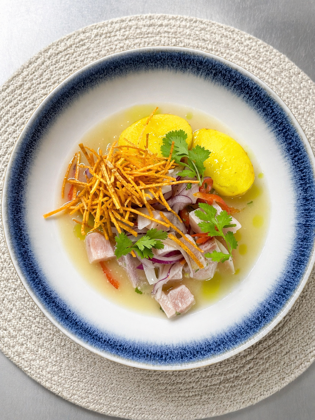 A bowl of ceviche with chopped fish, red onions, cilantro, sliced red chili, crispy plantain chips, yellow potato slices, and lime on a white dish with a blue rim, placed on a woven placemat.