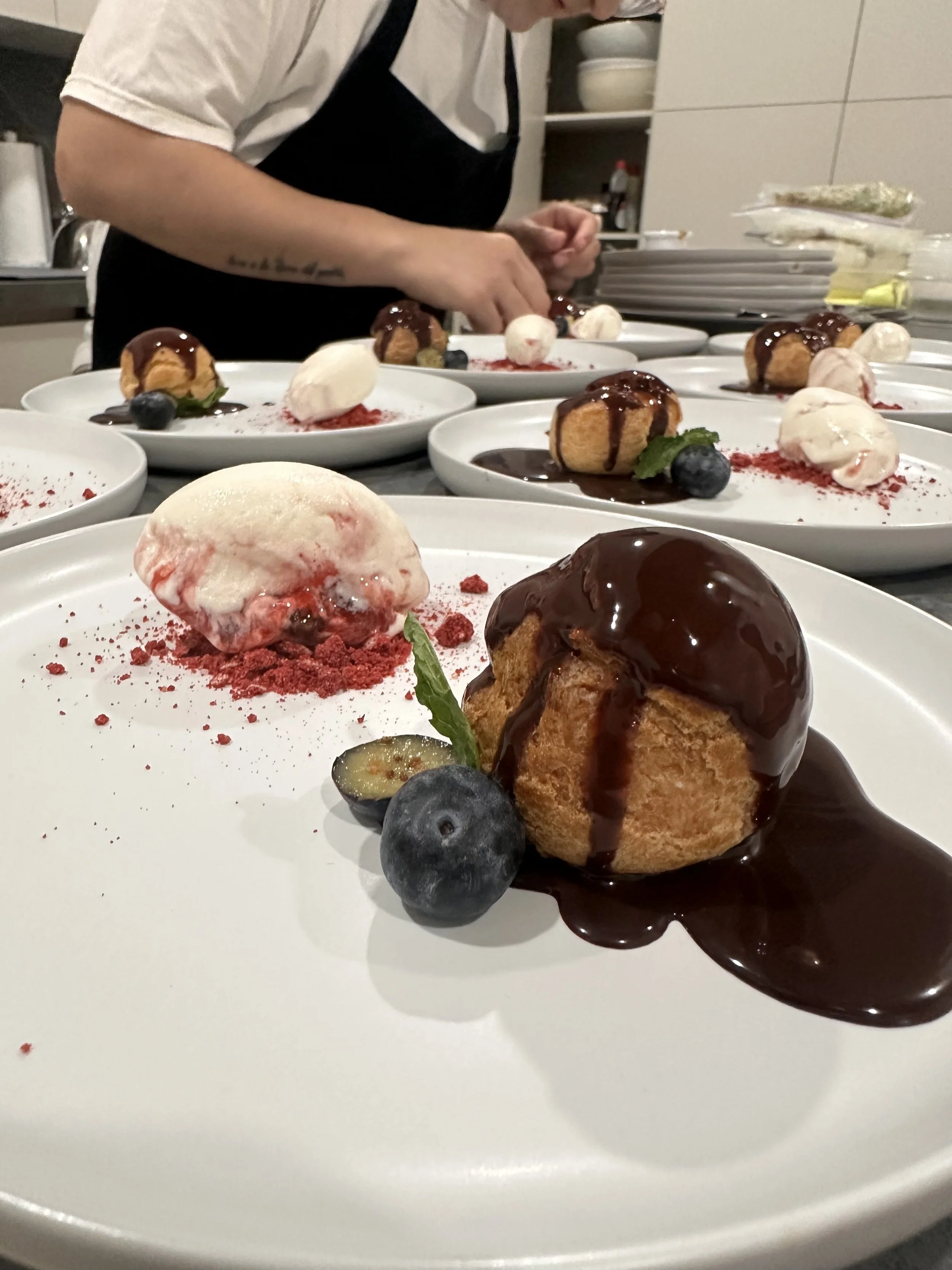 Multiple plates with dessert including profiteroles topped with chocolate sauce, some with and some without ice cream and berries, garnished with mint leaves.