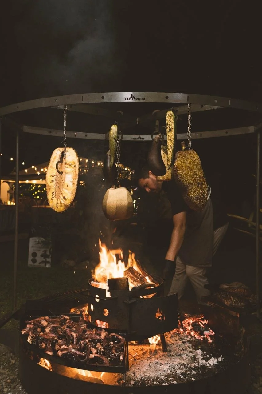 A person grilling meat over an open fire at night, with hanging dried and fresh fruit or vegetables above the fire, in an outdoor setting with string lights in the background.