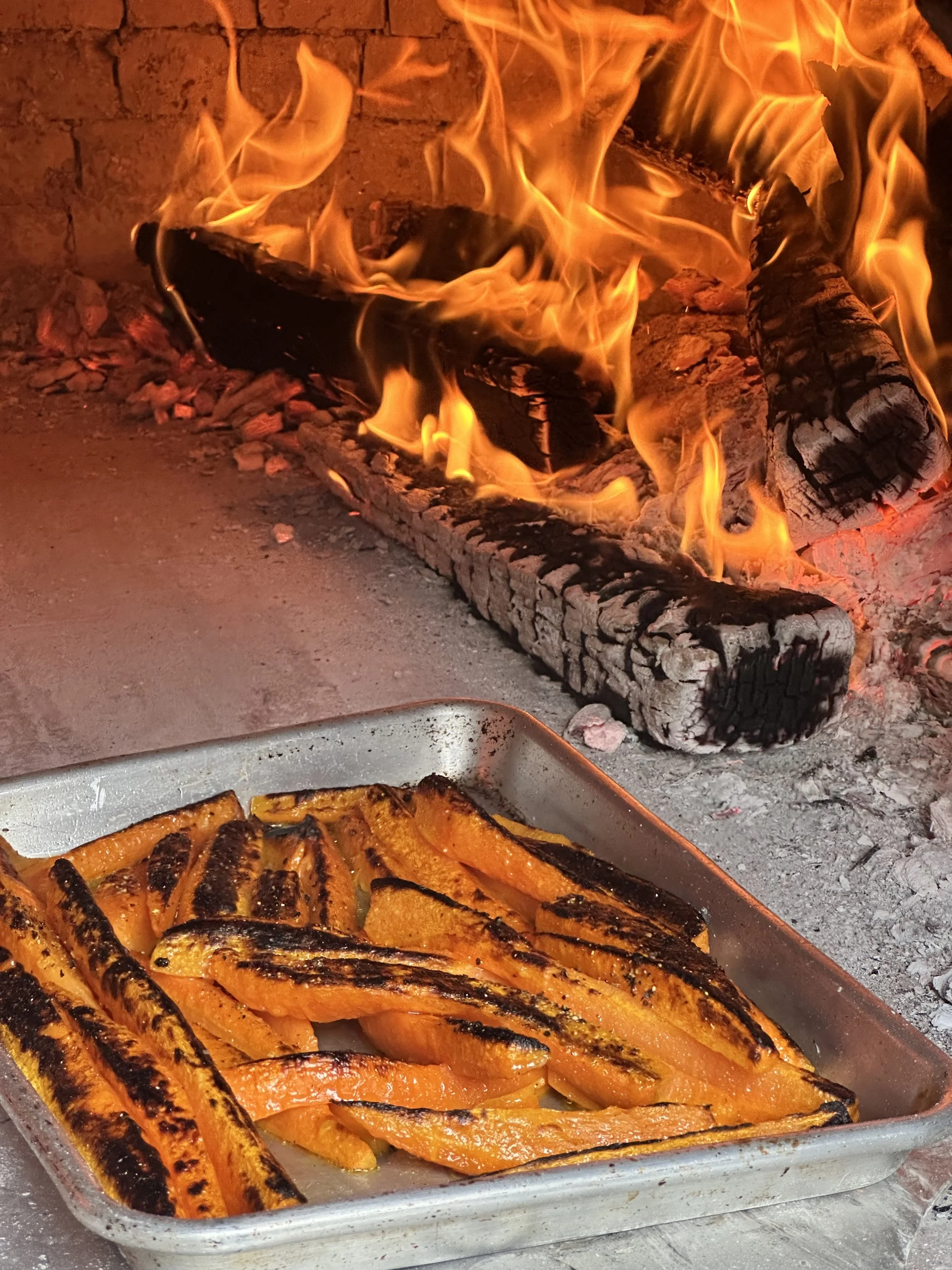 Charred grilled salmon and roasted orange slices on a metal tray in front of a brick fireplace with burning logs.