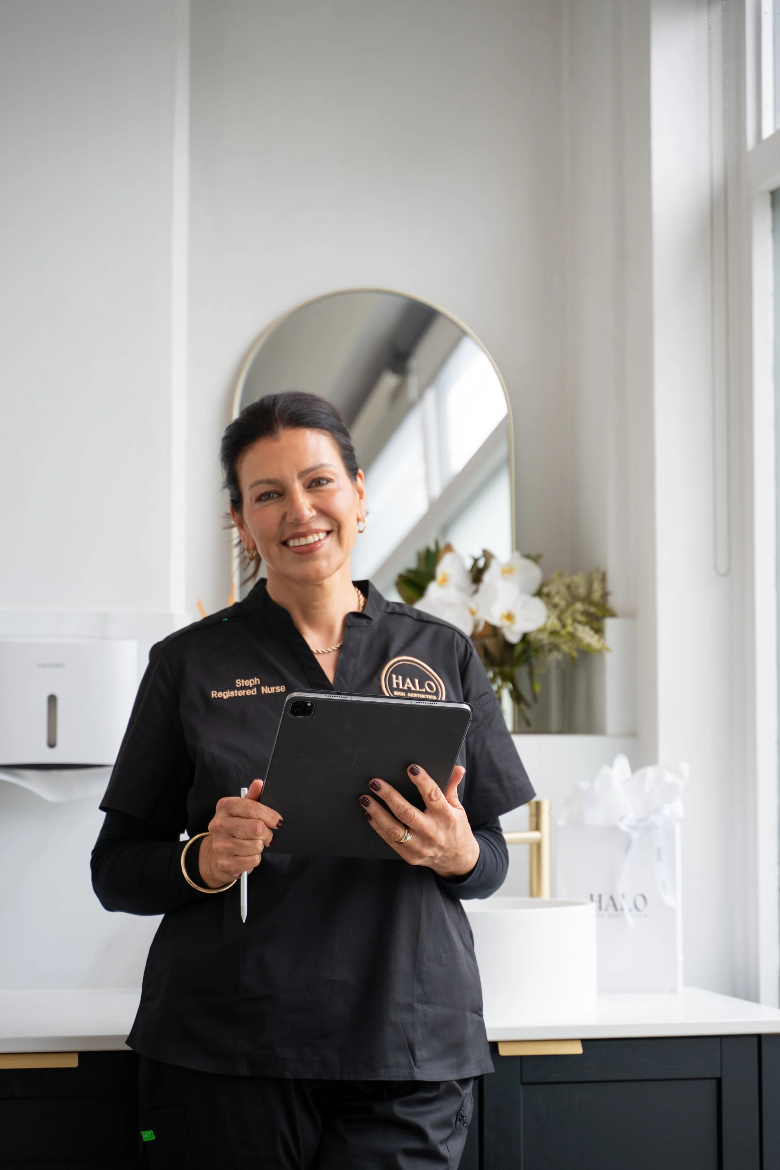 A smiling woman in black scrubs holds a tablet device indoors. She has dark hair and wears jewelry. Behind her is a large mirror, floral decoration, and a branded shopping bag in a minimal, modern setting.