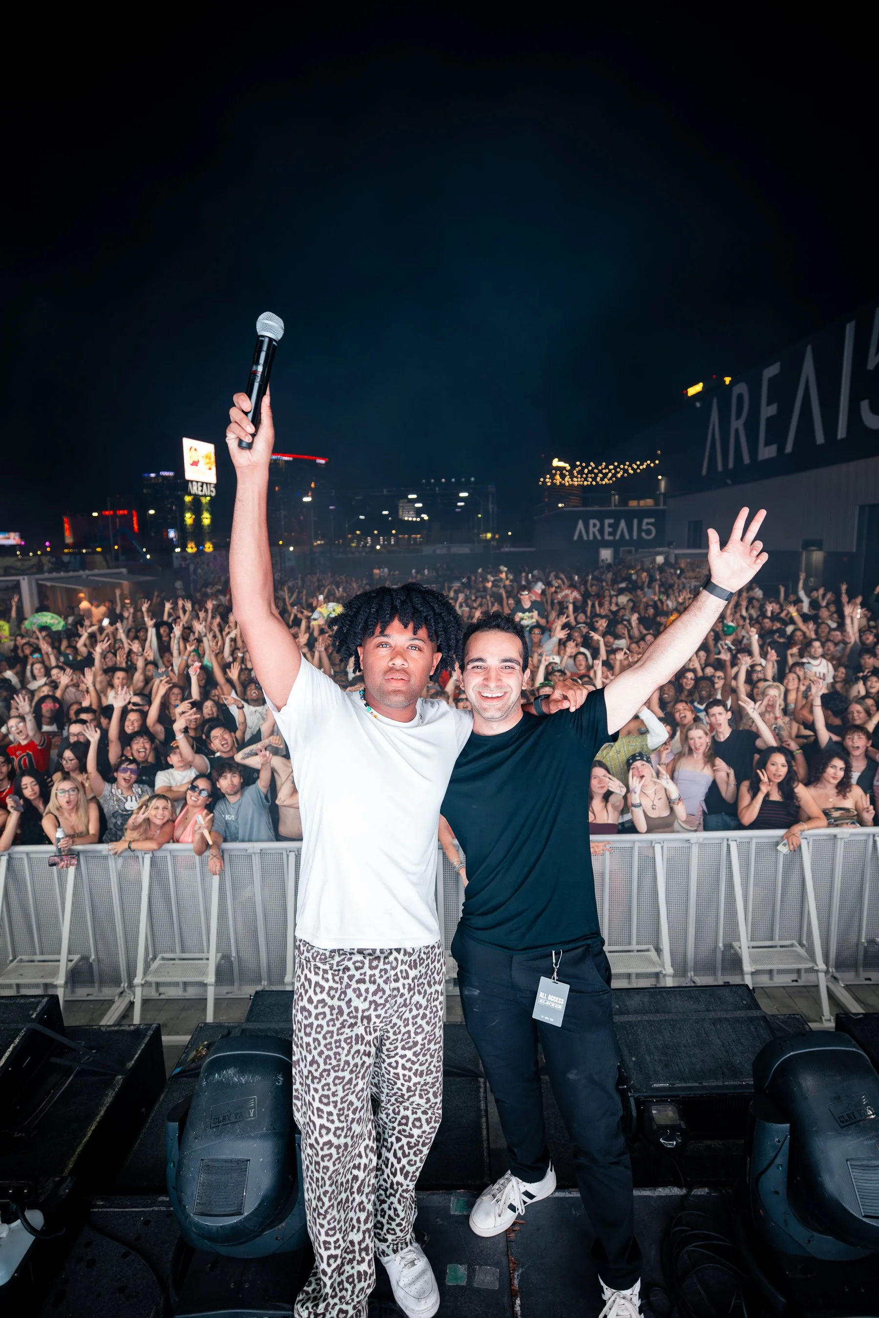 Two performers on stage in front of a large dance crowd at a music concert, one holding a microphone and raising their arm, empty night sky, illuminated cityscape, and concert signage in background.