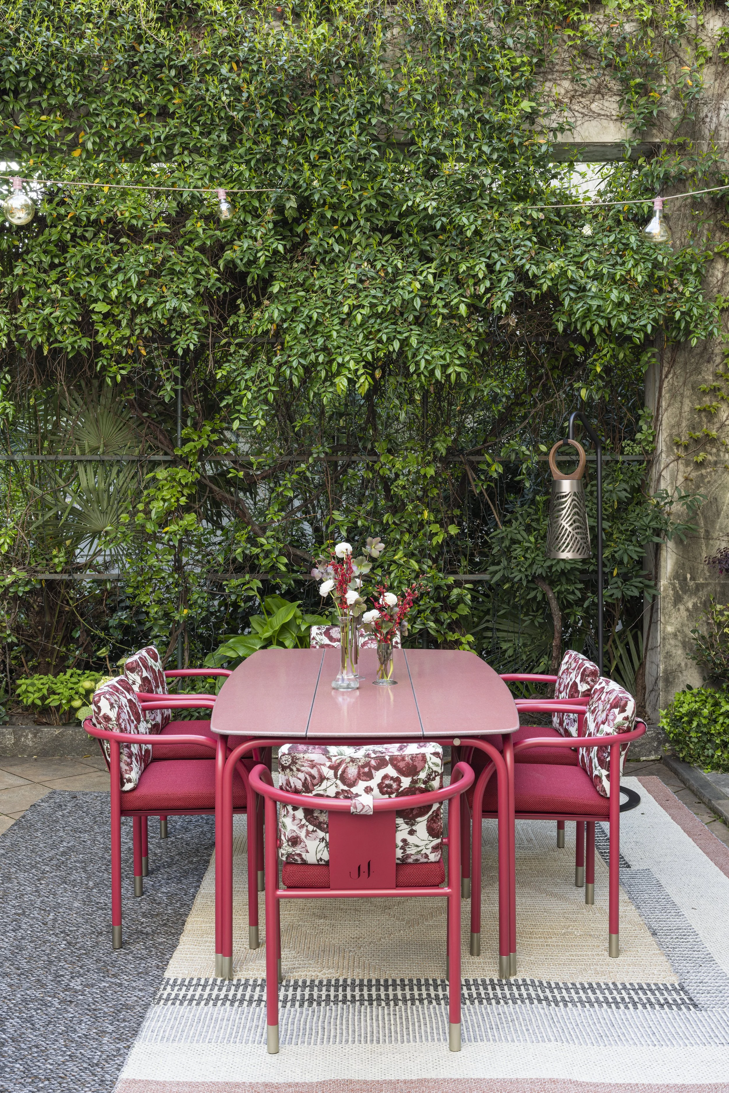 Tavolo rosa con sedie imbottite con motivo floreale, decorato con vasi di fiori, in un cortile con piante verdi e luci a festa.