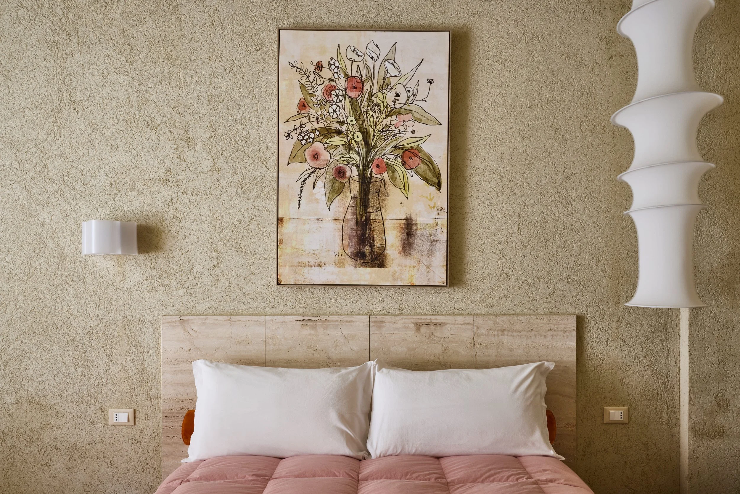 Camera da letto con pareti beige, quadretto con vaso di fiori sulla testiera, letto con lenzuola rosa e cuscini bianchi, lampada da parete e lampada decorativa bianca verticale.