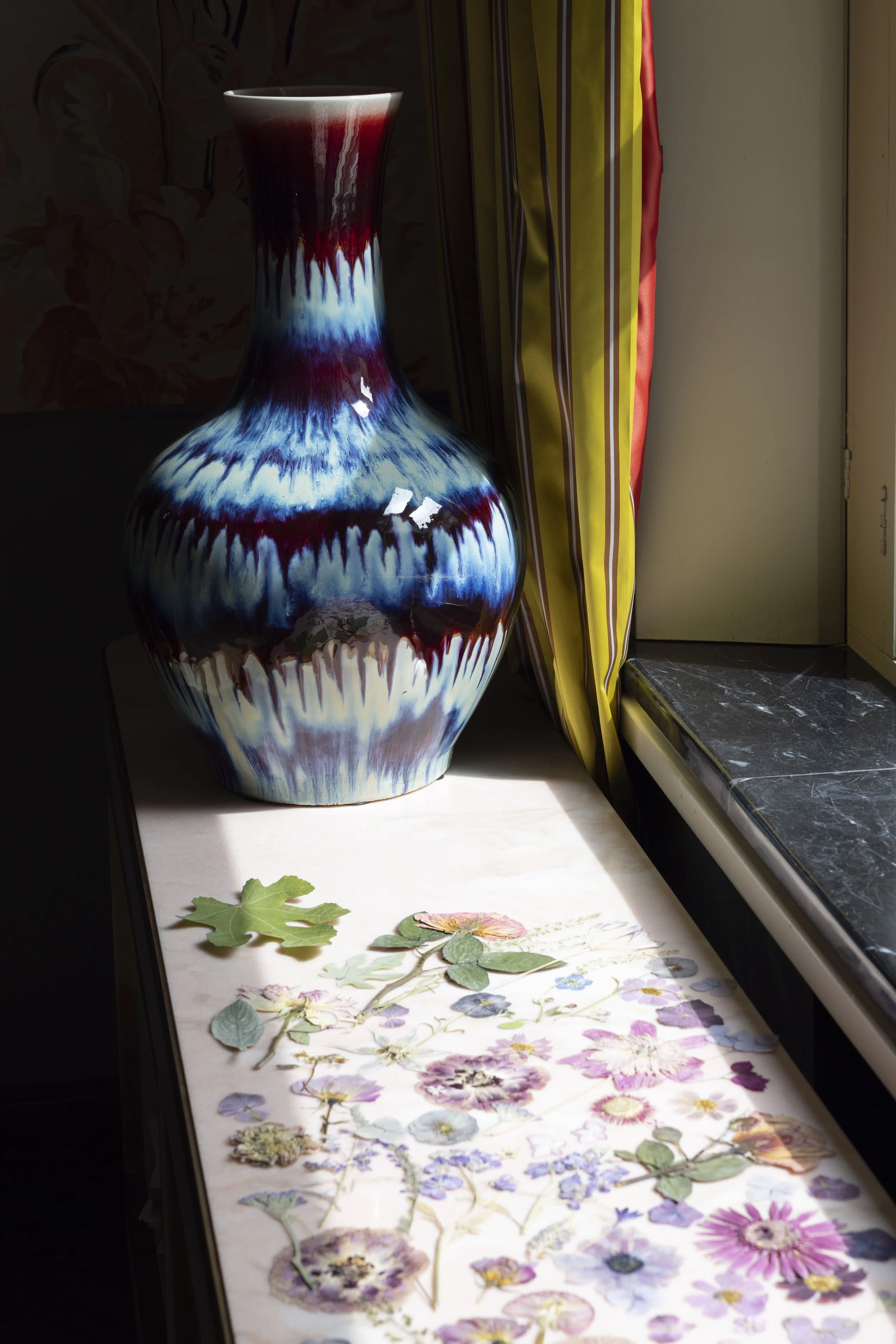 Vaso di ceramica con decorazioni colorate su una finestra, accanto a tenda gialla con strisce rosse e verdi, sopra un tavolo con foglie e fiori secchi