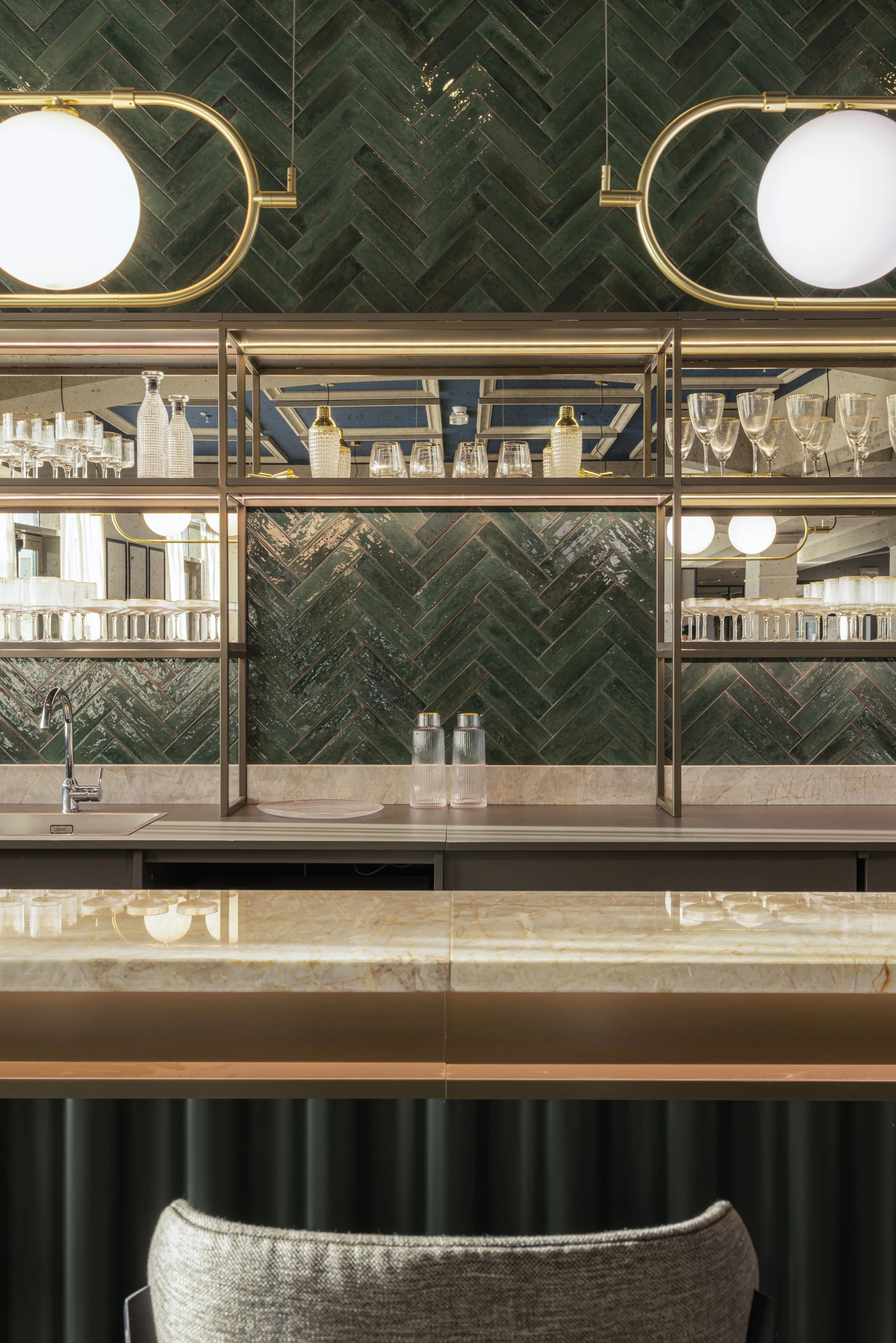 Interno di un bar o ristorante elegante con scaffali di vetro con bicchieri e bottiglie, pareti di piastrelle verdi in stile a spina di pesce, e luci da tavolo a forma di globi.