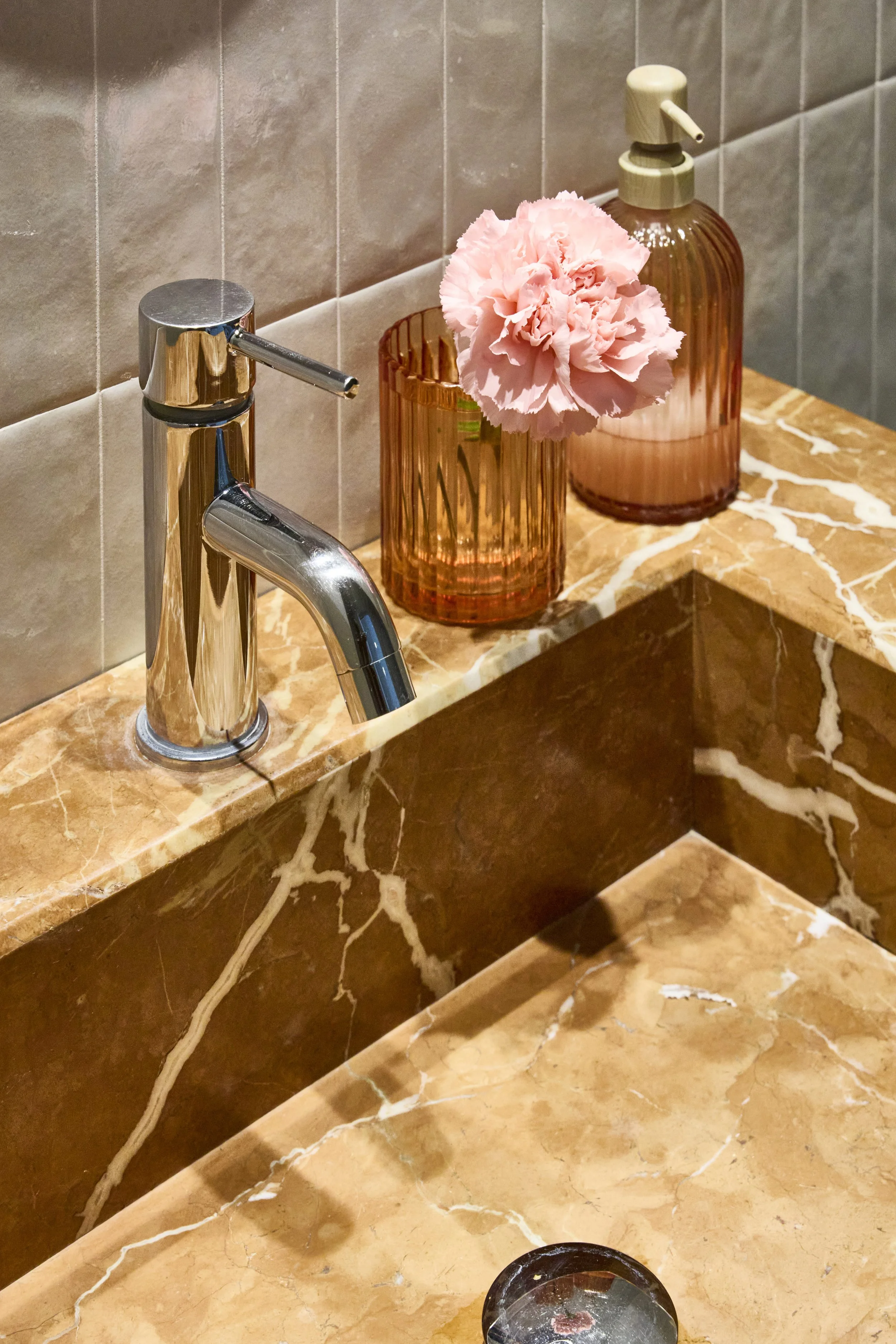 Lavandino con rubinetto cromato, due vasi di vetro rosa con un fiore rosa, sapone e un portascopino, in un bagno con pareti in piastrelle beige.