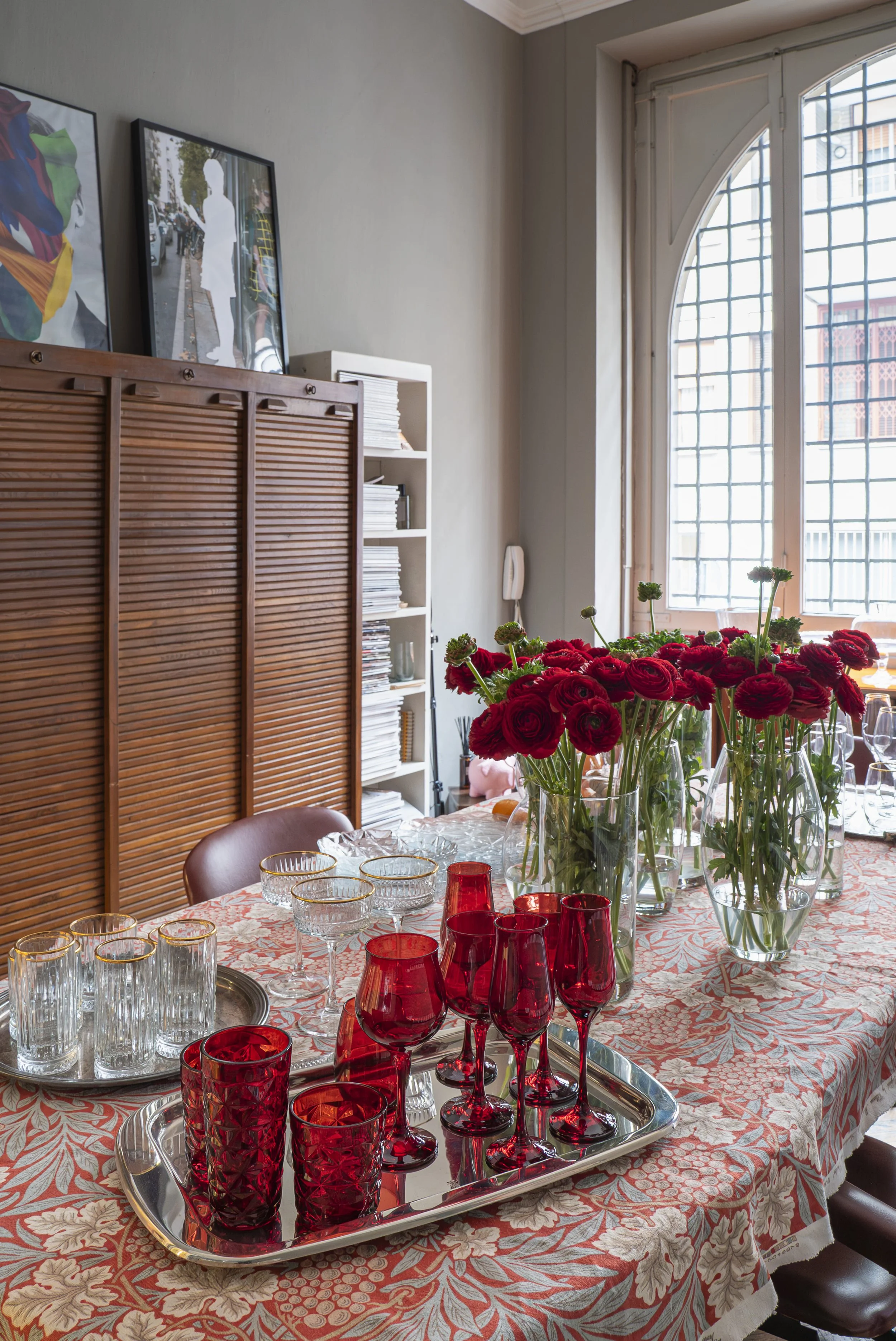 Tavolo decorato con fiori rossi in vasi di vetro e bicchieri di cristallo, in una stanza luminosa con grande finestra.