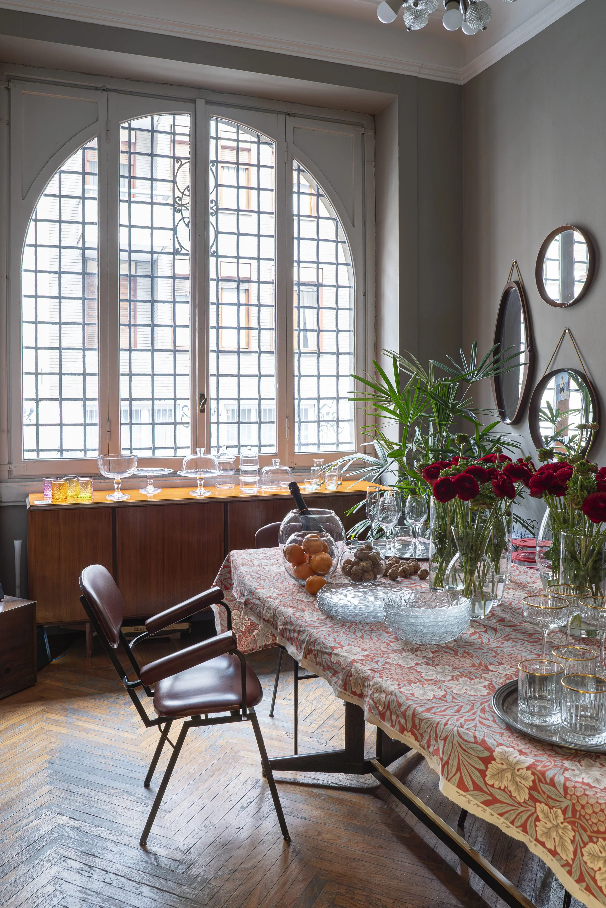 Sala da pranzo con grande finestra, tavolo decorato con fiori e bicchieri di vetro, pareti decorate con specchi rotondi, piante verdi, sedie in pelle marrone, pavimento in parquet.