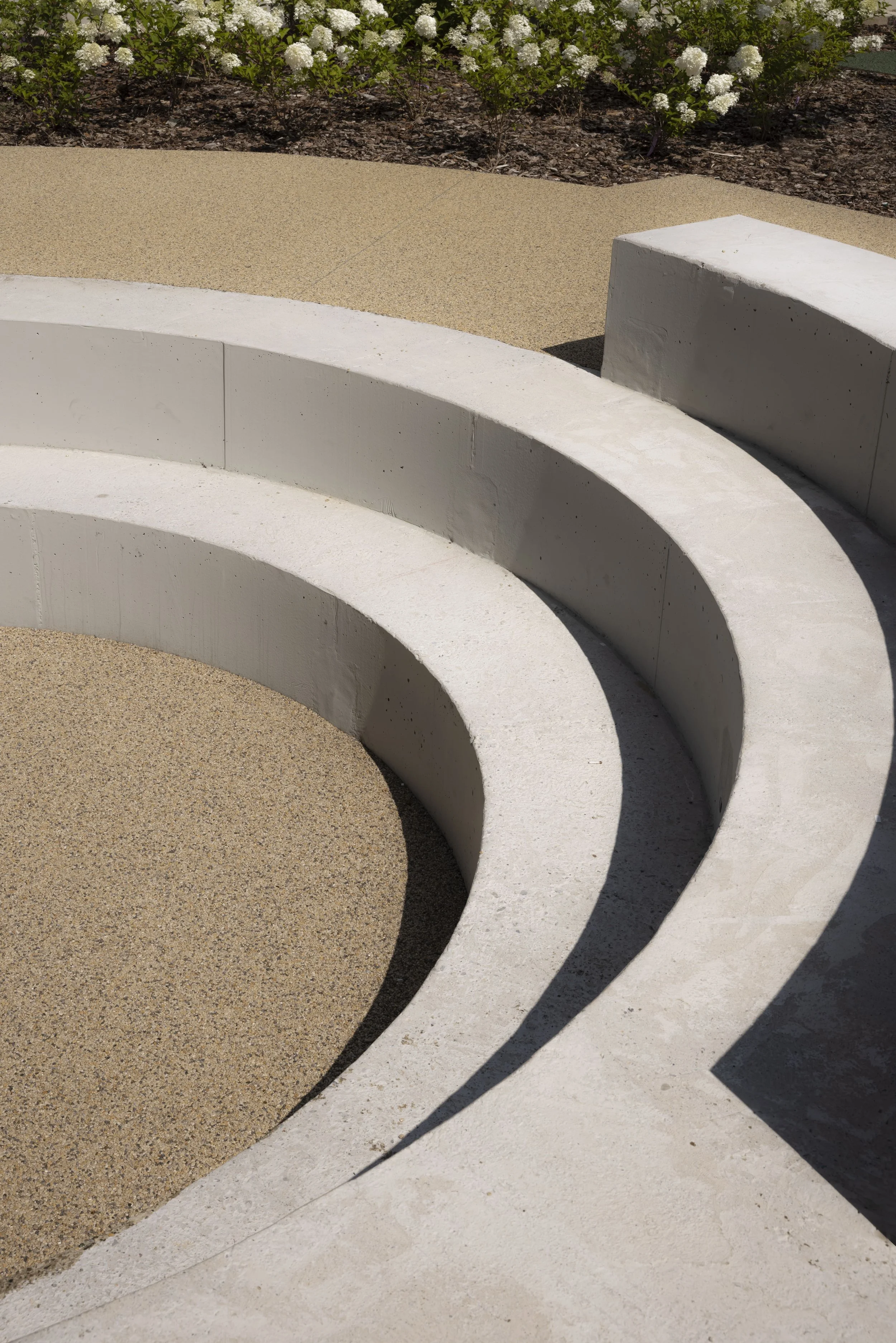 Seduta circolare in cemento bianco con superficie di sabbia o ghiaia, con fiori bianchi e piante verdi sullo sfondo.