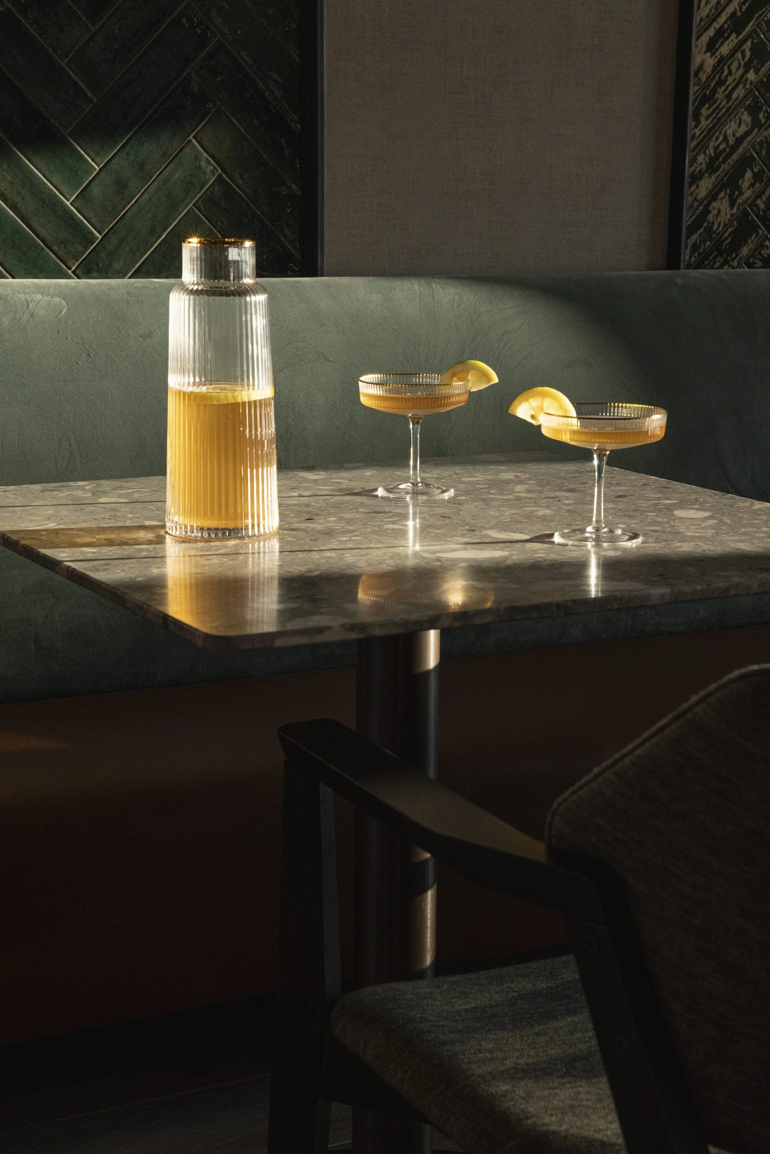 Una bottiglia di vetro con una bevanda gialla e due cocktail con fette di limone sono posizionati su un tavolo di marmo in un ambiente elegante, con arredi scuri e luci soffuse.