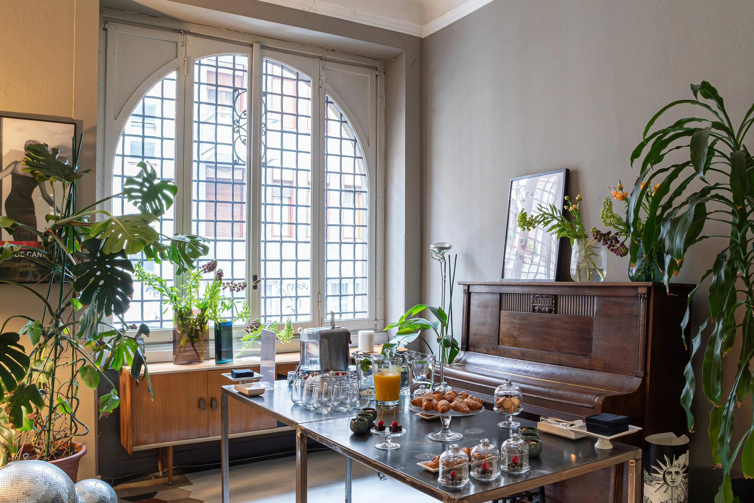 Sala da pranzo con grandi vasi di piante verdi, tavolo con bevande e dolci, finestra grande con grate e luce naturale, mobili in legno, decorazioni floreali.