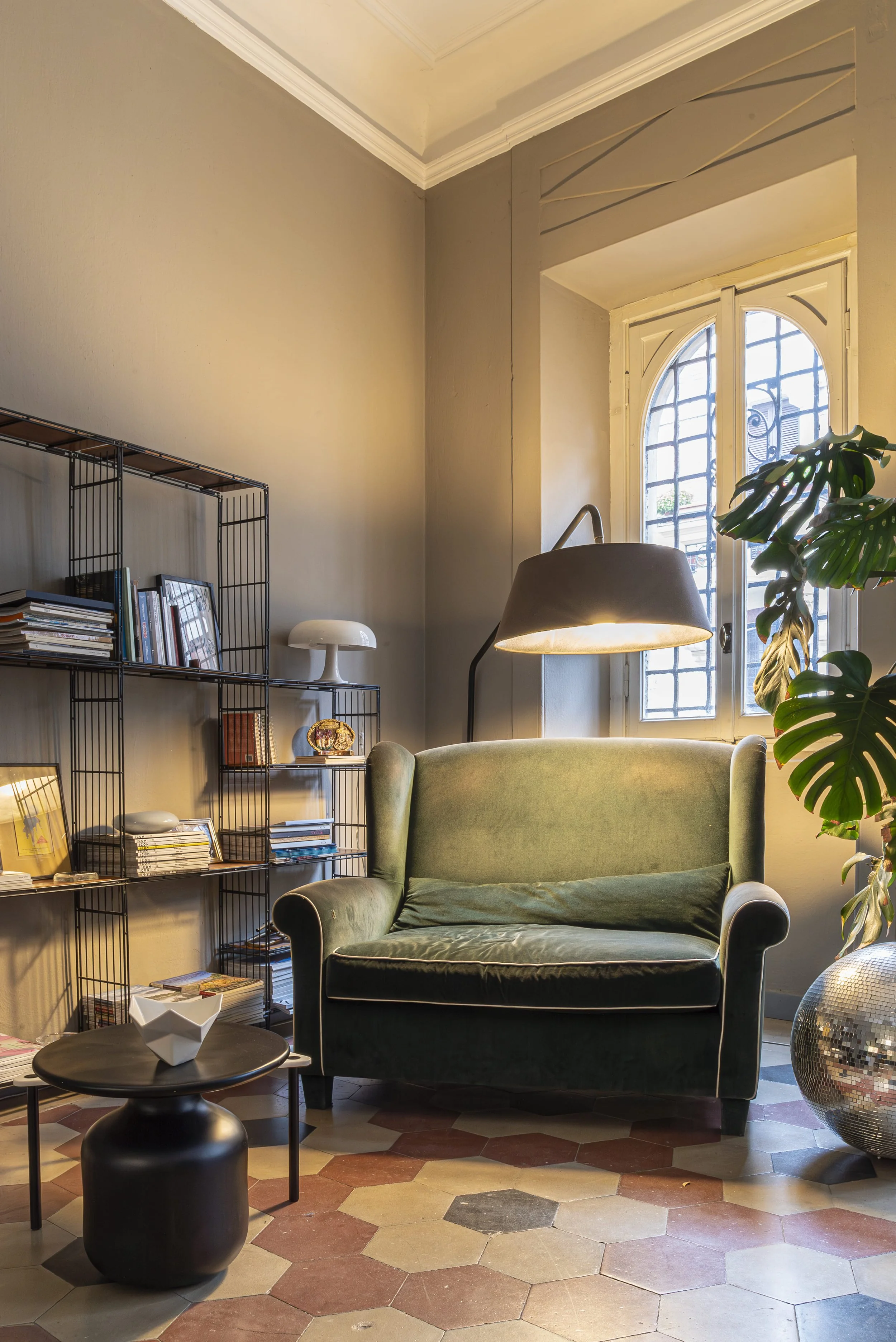 Interno: salotto con divano verde, libreria in metallo nera, lampada da terra e grande pianta tropicale.