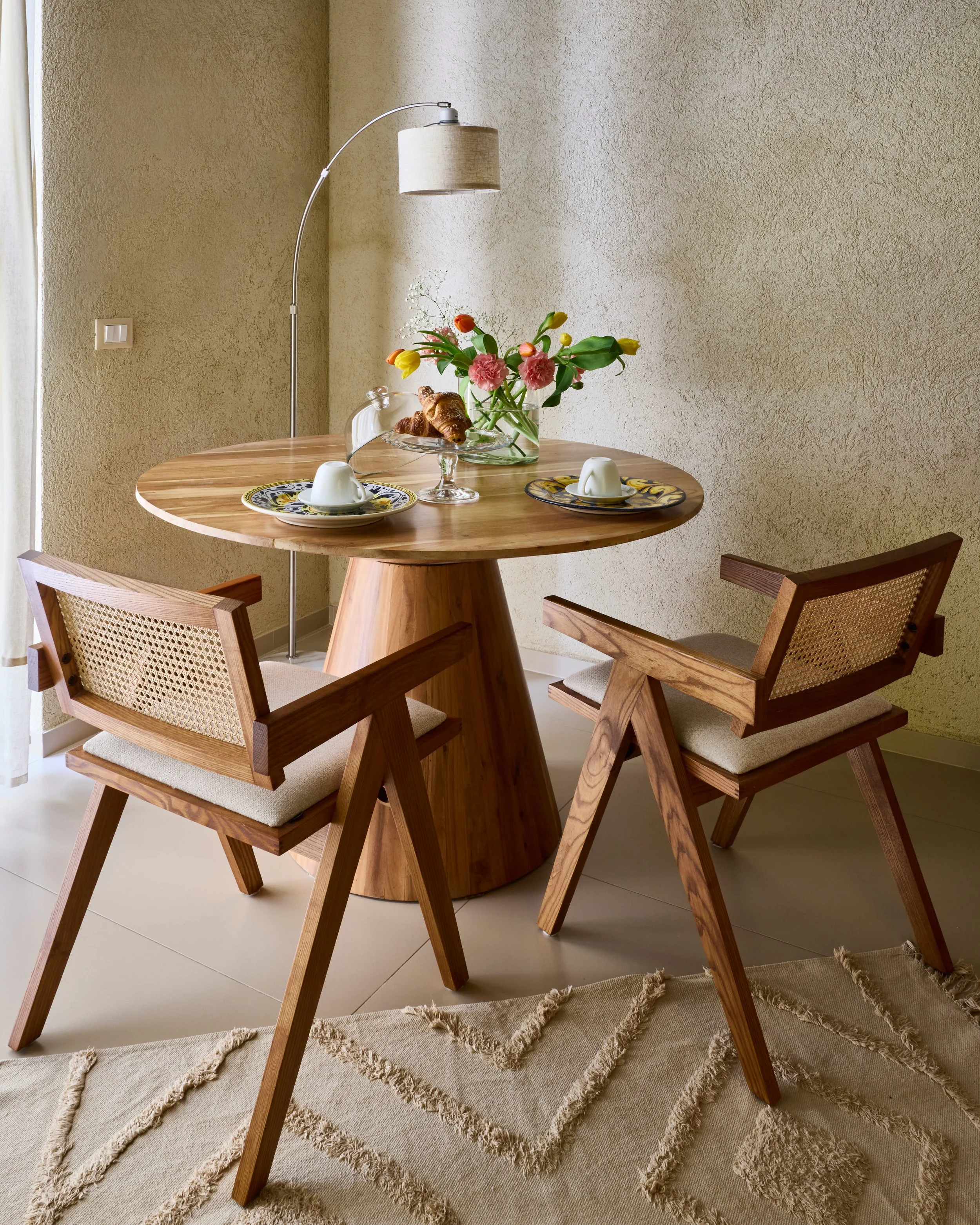 Tavolo rotondo in legno con due sedie in legno, centro tavola con fiori colorati, piatti con tazze, lampada da terra lunga e una parete beige