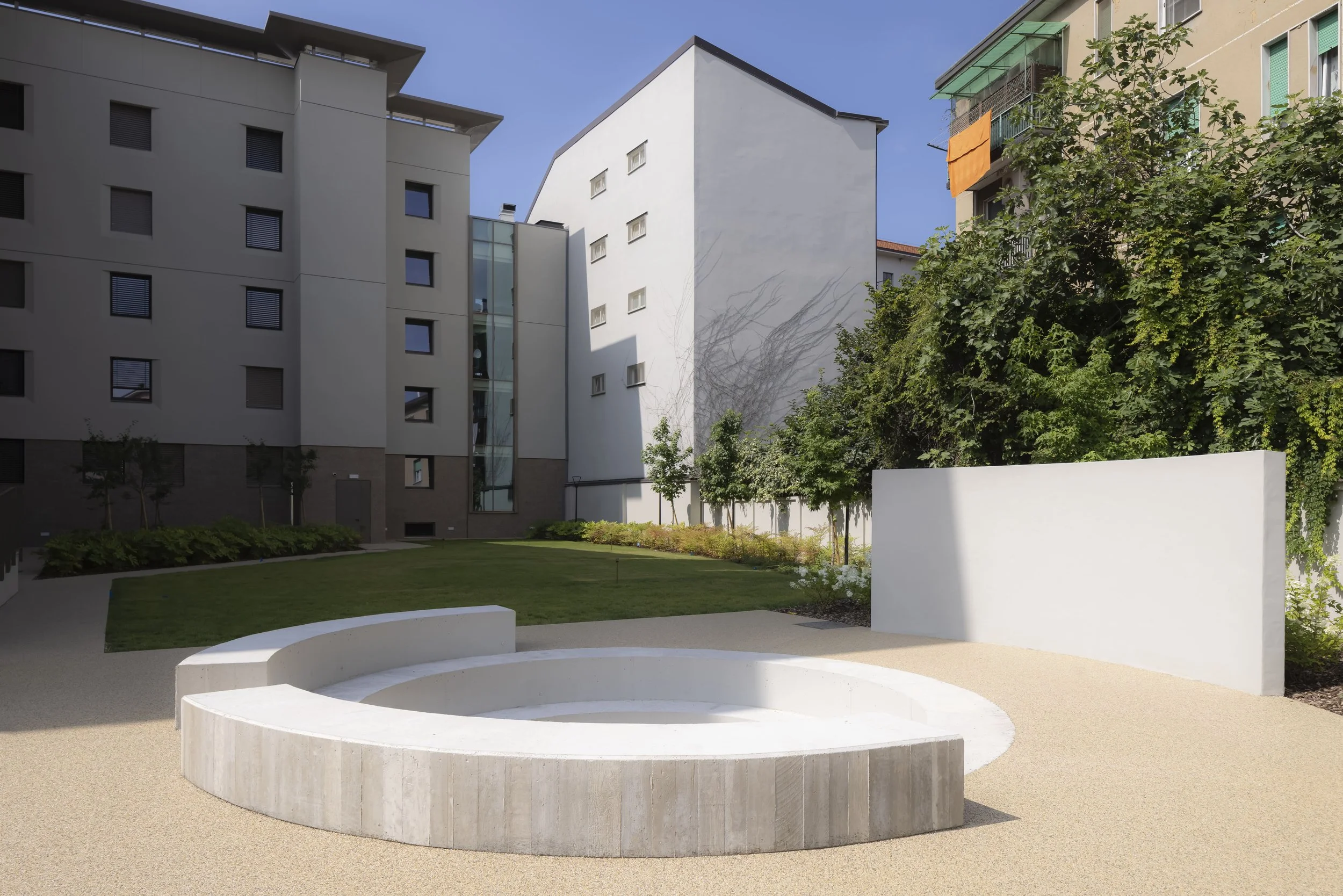 Giardino con area verde, panchina curva bianca in cemento e parete bianca tra edifici moderni, alberi e piante