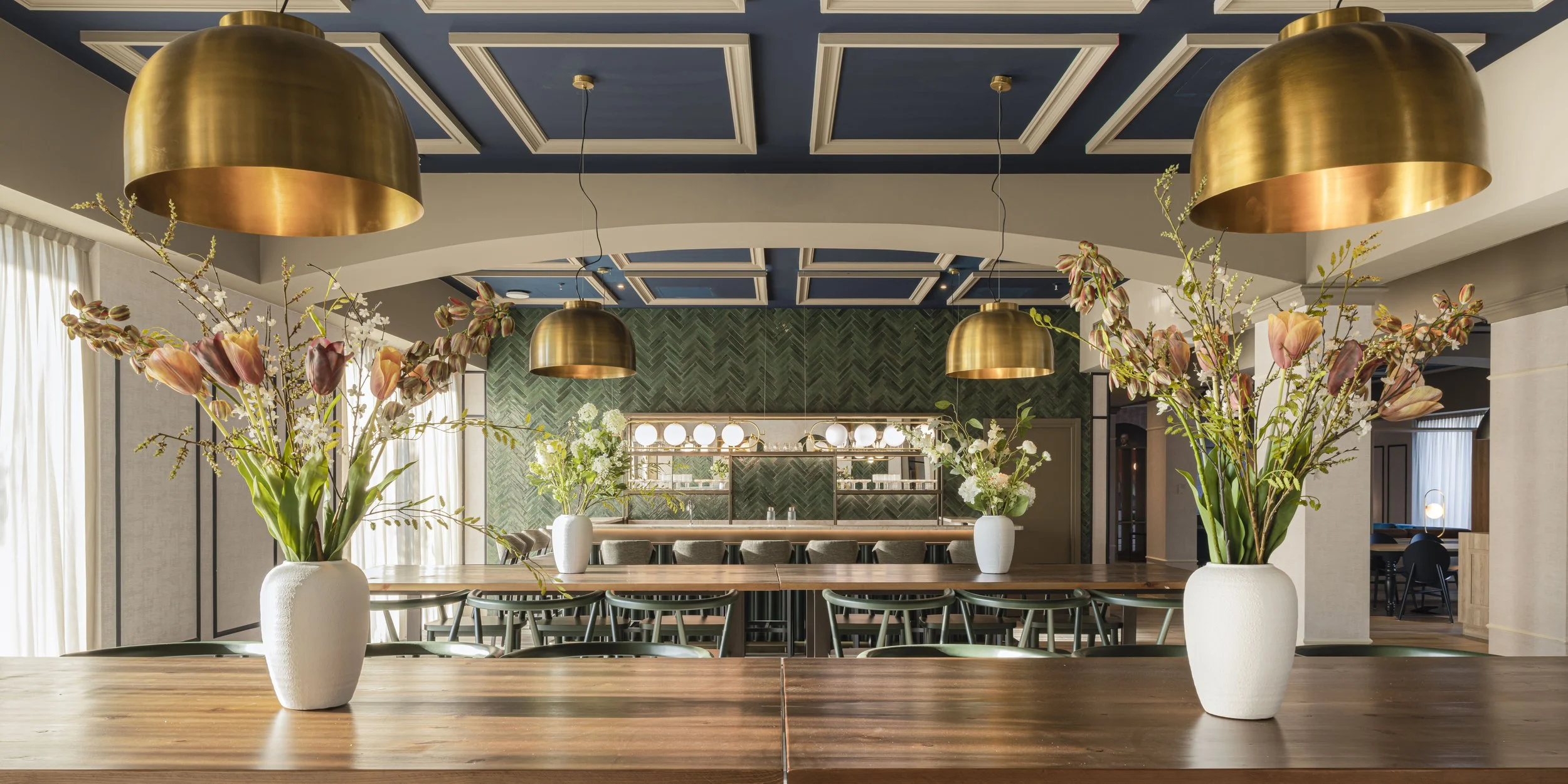 Interno di un ristorante elegante con tavoli in legno, grandi vasi con fiori e luci a sospensione dorate.