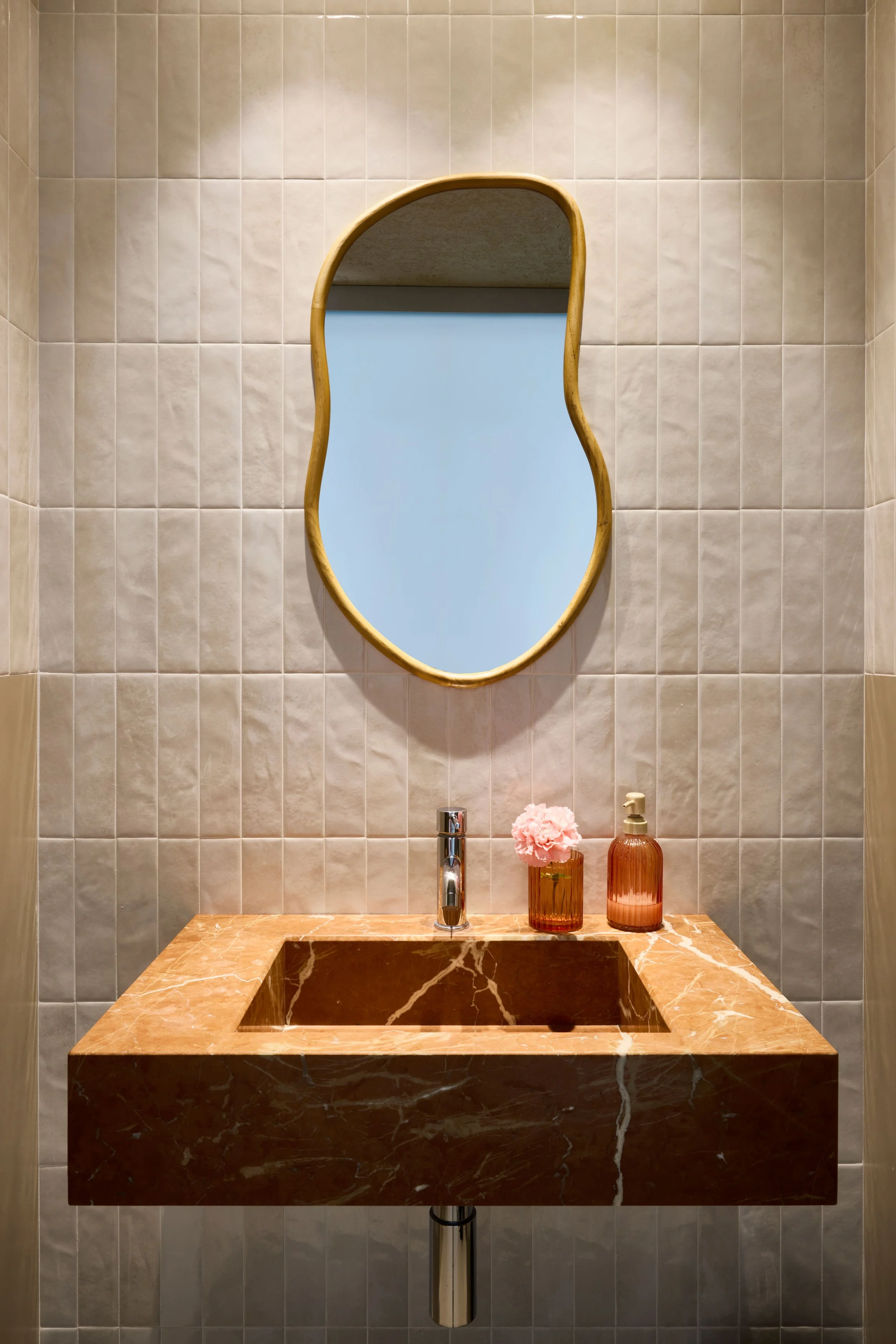 Specchio curvo sopra un lavabo di marmo rosso con un dispenser di sapone e un vaso con fiore rosa.