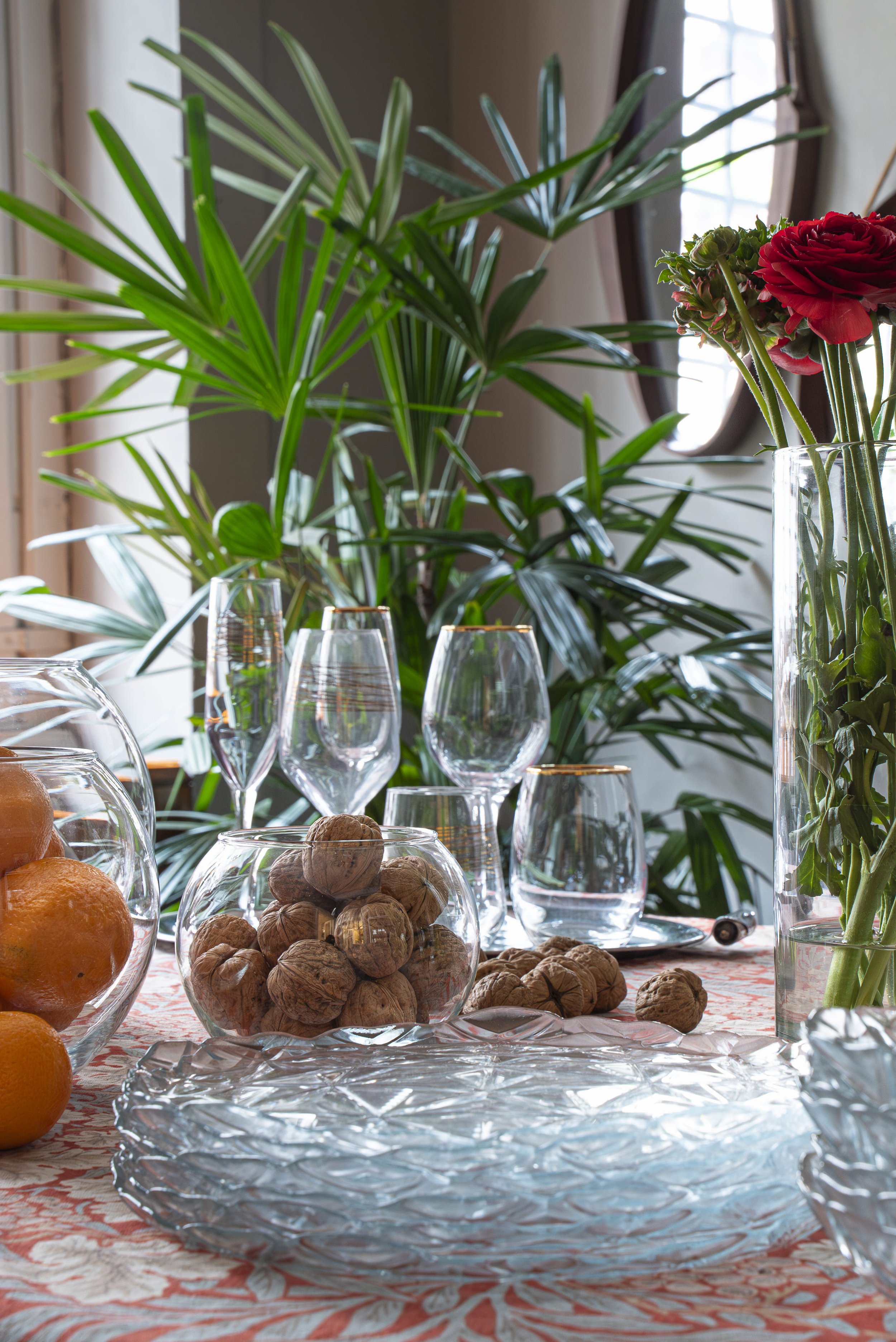 Tavolo apparecchiato con ciotole di noci e agrumi, bicchieri di vetro, un vaso con fiori rossi e un grande pianta verde in una stanza luminosa.