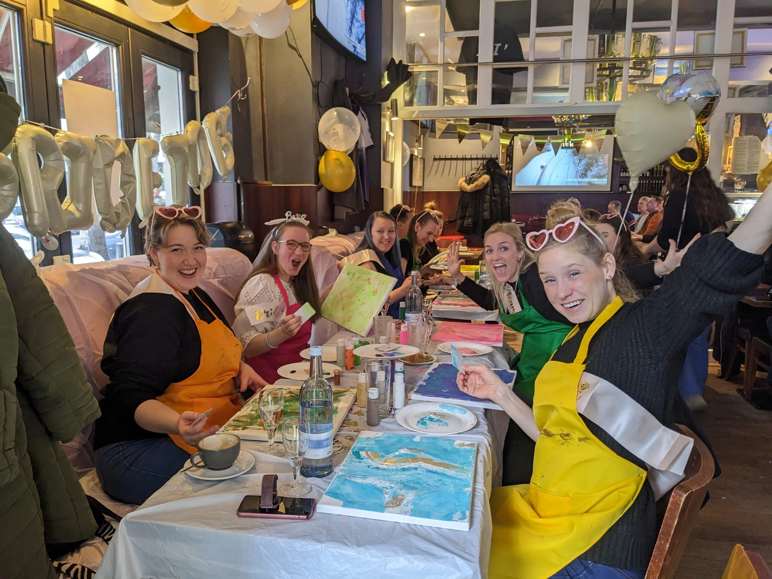 Gruppe von Frauen, die an einem Tisches in einem festlich dekorierten Raum malen und lachen, mit Ballons und Dekorationen im Hintergrund.