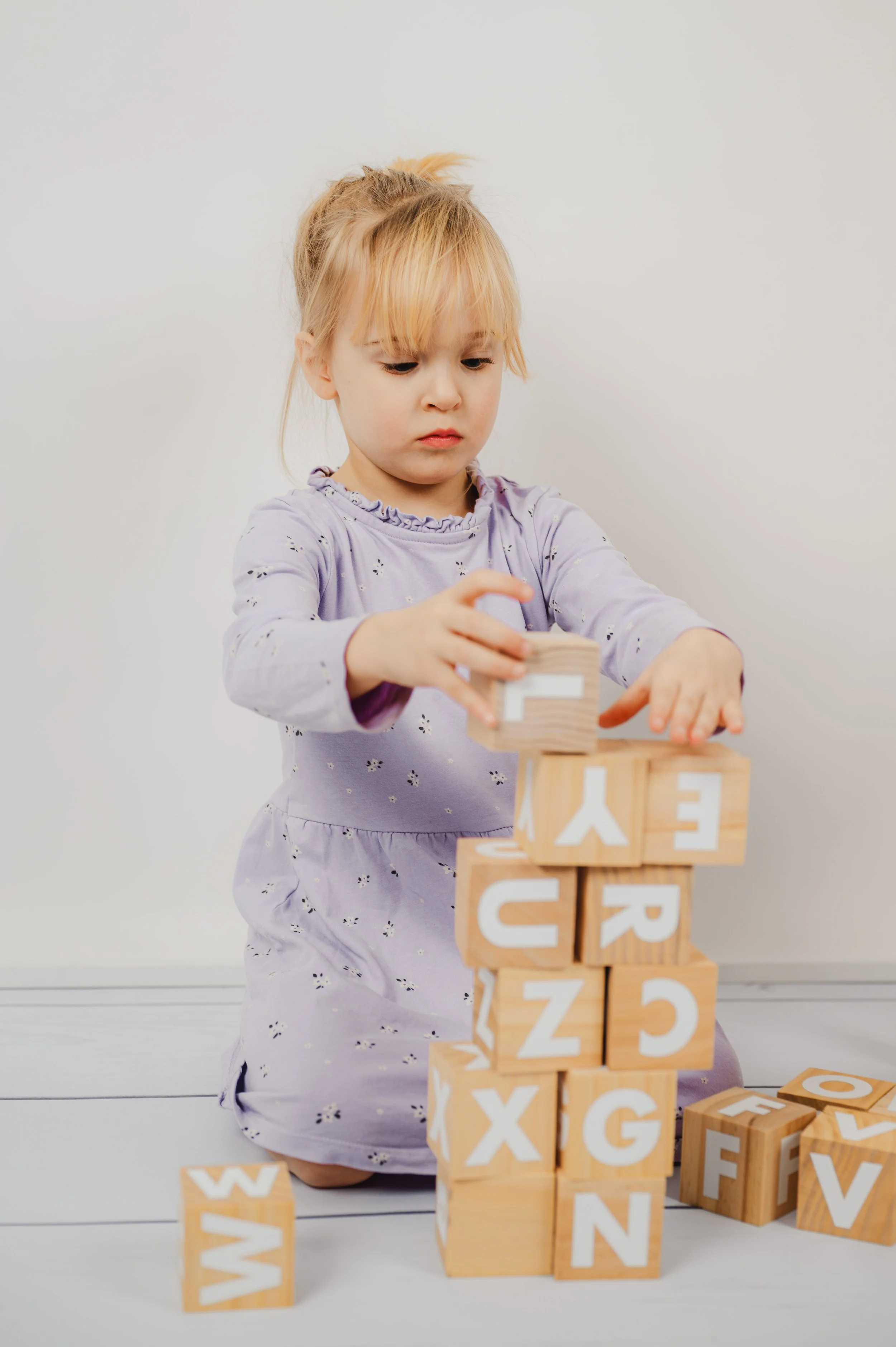Natürliches Kita- und Schulfoto eines Kindes beim Stapeln von Holzwürfeln. Spielerische Szene mit Holzbuchstaben vor hellem, ruhigem Hintergrund. Authentische, ungezwungene Schulfotografie mit Fokus auf Konzentration, Motorik und kindliches Spiel.