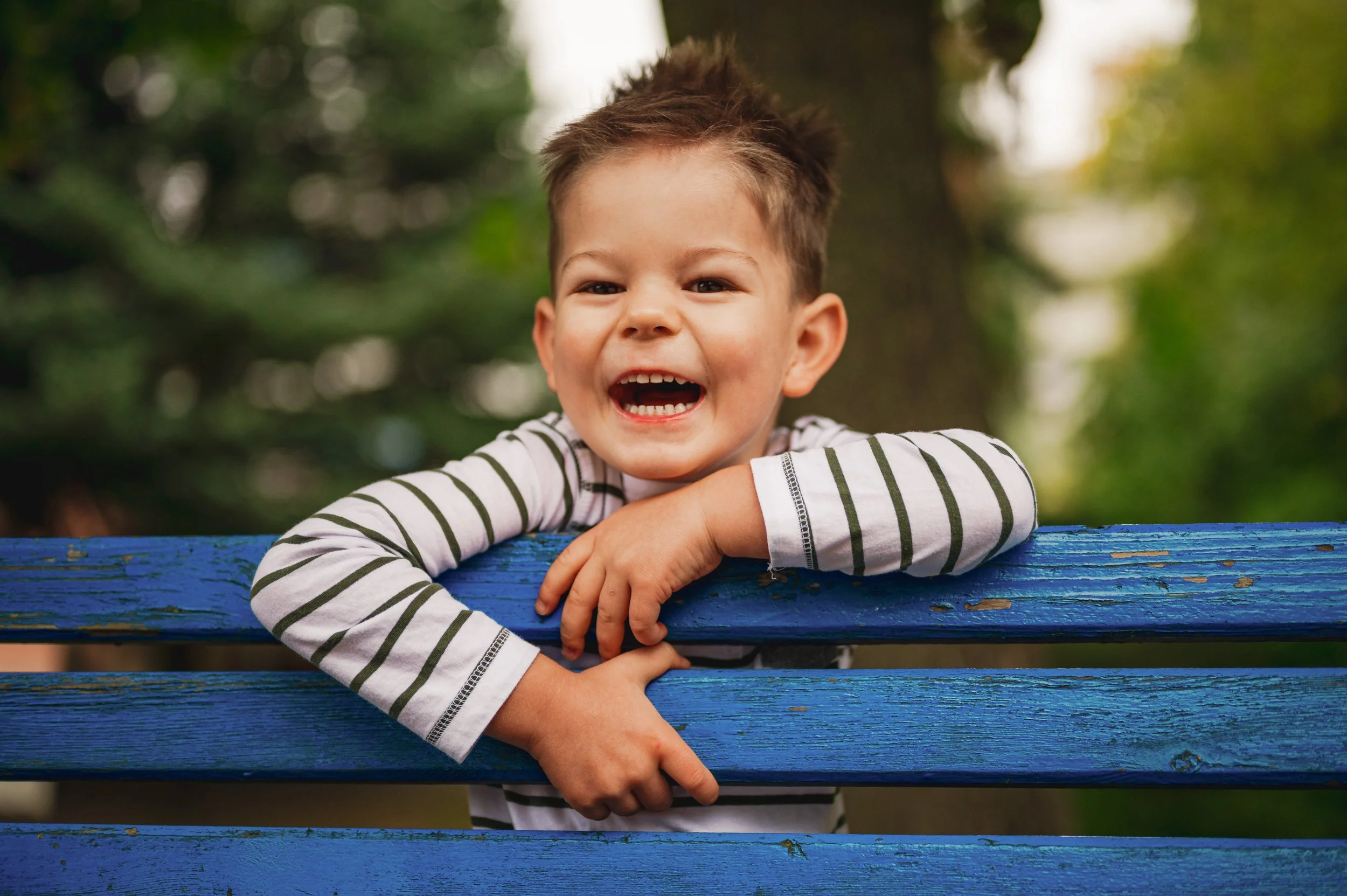 Fröhliches Kita-Kind lehnt lachend an einer blauen Bank im Grünen – natürliche Kita-Fotografie in Sachsen und Brandenburg von KUNTERBUNTE MOMENTE.