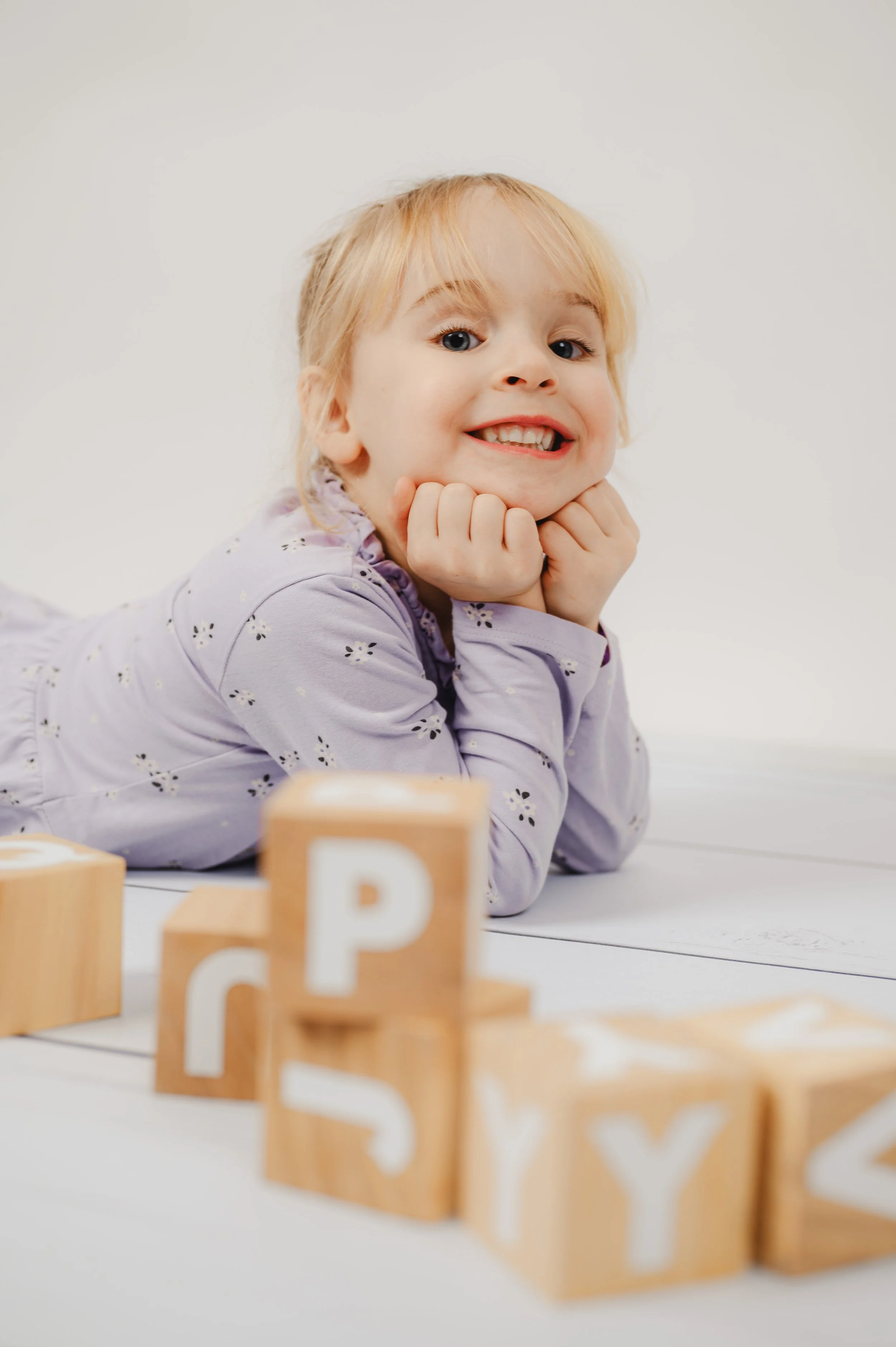 Natürliches Kita- und Schulfoto eines Kindes, das entspannt vor Holzwürfeln liegt und fröhlich in die Kamera lächelt. Ruhiger, heller Hintergrund und spielerische Holzbuchstaben schaffen eine warme, authentische Atmosphäre. Moderne Kita-Fotografie mi