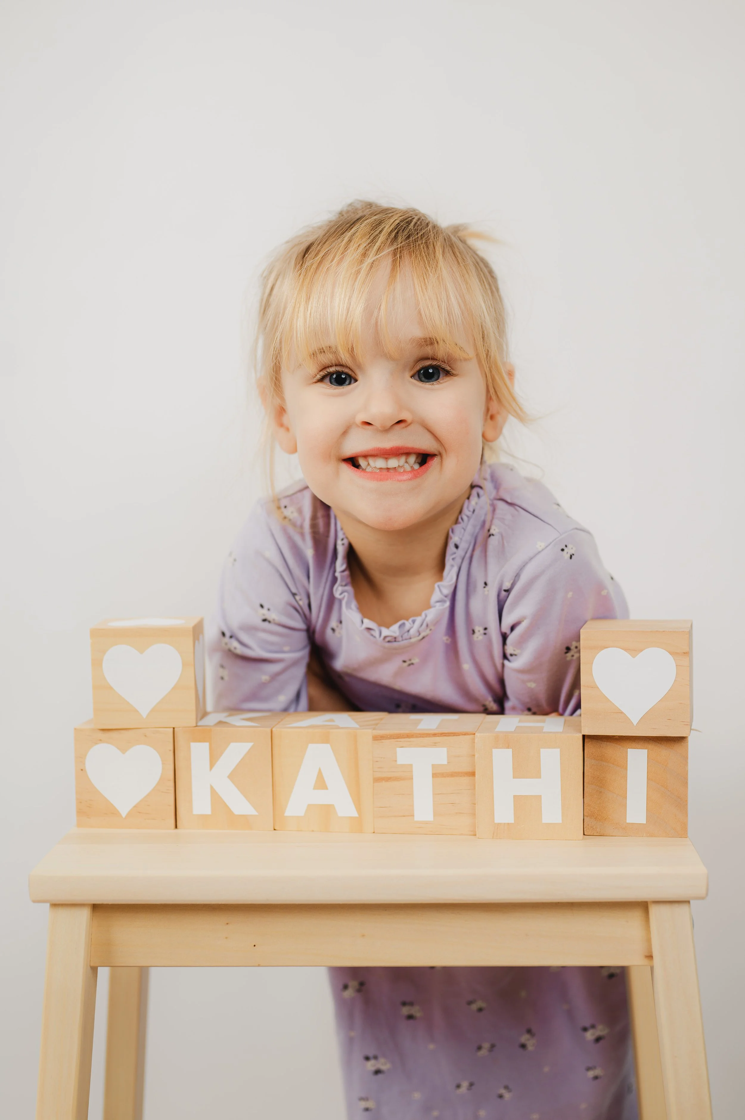 Natürliches Kita- und Schulfoto eines Kindes, das fröhlich hinter Holzwürfeln steht und in die Kamera lächelt. Die Holzklötze sind zu einem Namen gestapelt und schaffen eine spielerische, persönliche Szene. Heller Hintergrund, ruhige Farben und echte