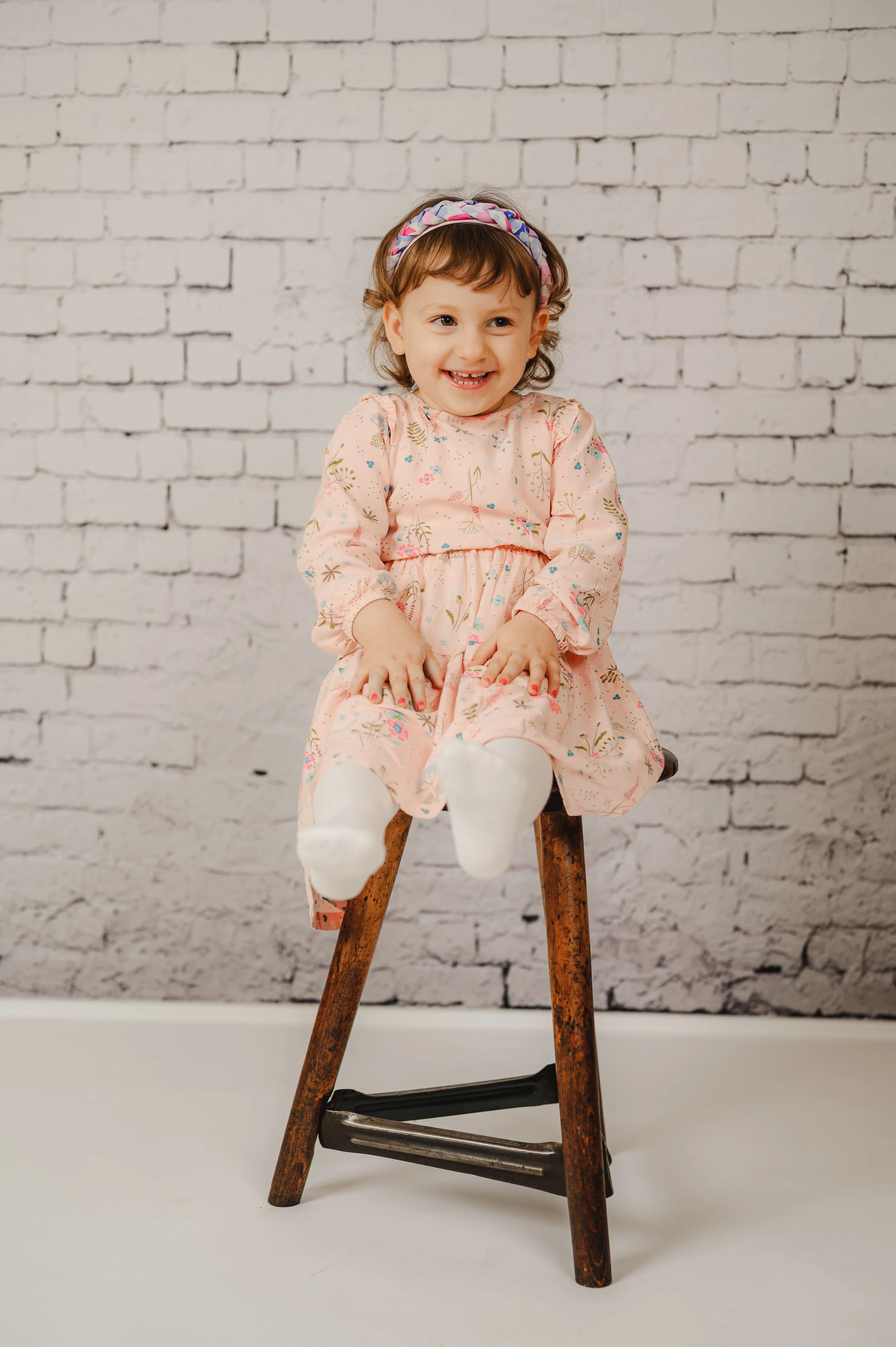 Ein fröhliches Kinderporträt im Kreidetafel-Set:
Das Kind sitzt entspannt auf einem alten Werkstatthocker vor einer hellen Backsteinwand und lacht ganz natürlich in die Kamera.
Die ruhige Studioatmosphäre, dezente Farben und reduziertes Setting lenke