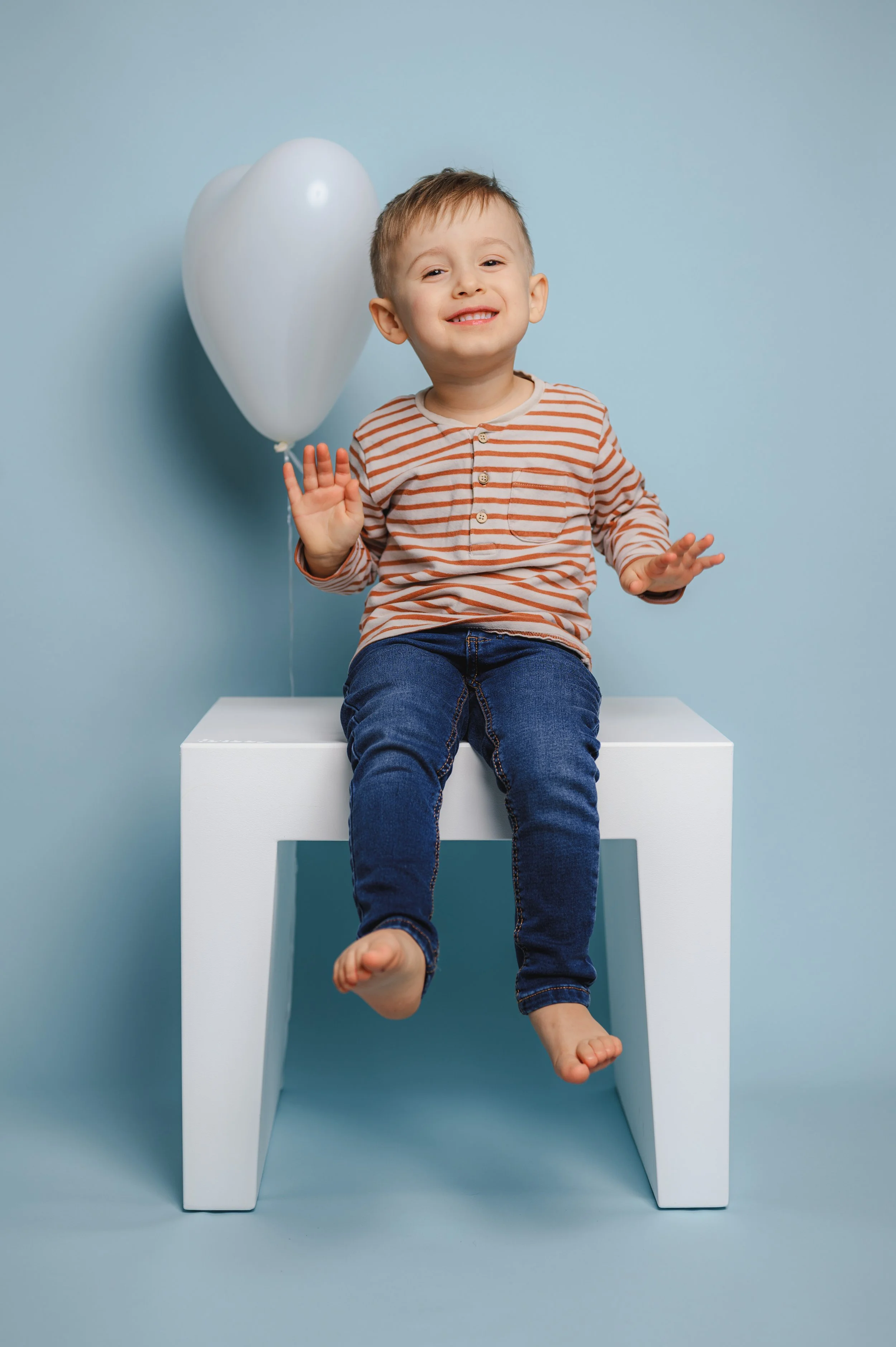 Natürliches Kita-Foto eines fröhlichen Kindes, das locker auf einem weißen Hocker sitzt. Ein weißer Luftballon und der ruhige blaue Hintergrund schaffen eine helle, freundliche Atmosphäre für moderne und ungezwungene Kita-Porträts.