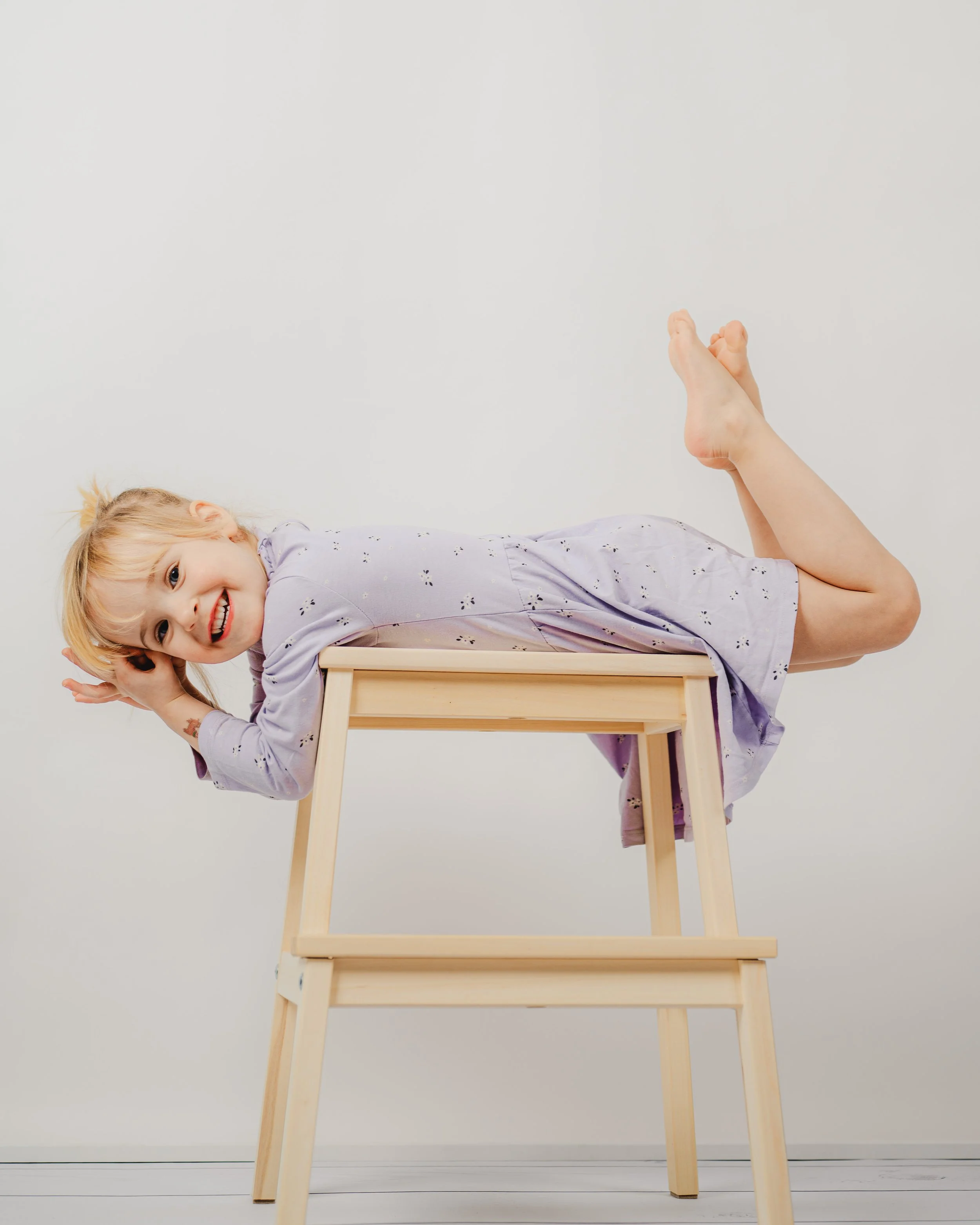 Natürliches Kita- und Schulfoto eines Kindes, das entspannt und verspielt auf einem Holz­hocker liegt. Die lockere Pose, der helle Hintergrund und die ruhige Farbwelt sorgen für eine moderne, zeitlose Bildwirkung. Authentische Momente, echte Freude u