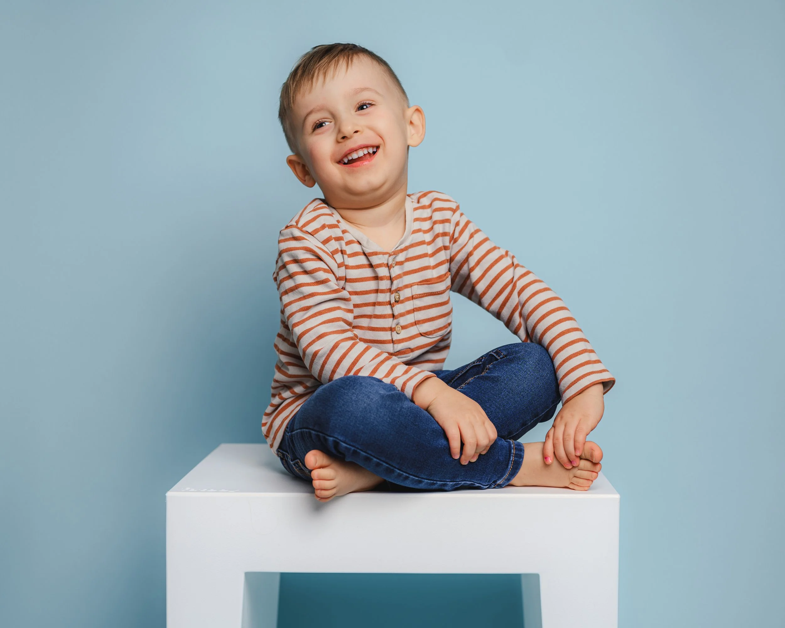 Fröhliches Kita-Foto eines lachenden Kindes, das entspannt auf einem weißen Hocker vor blauem Hintergrund sitzt. Das minimalistische Luftballon-Set sorgt für eine helle, freundliche Stimmung und eignet sich ideal für natürliche und lebendige Kita-Por