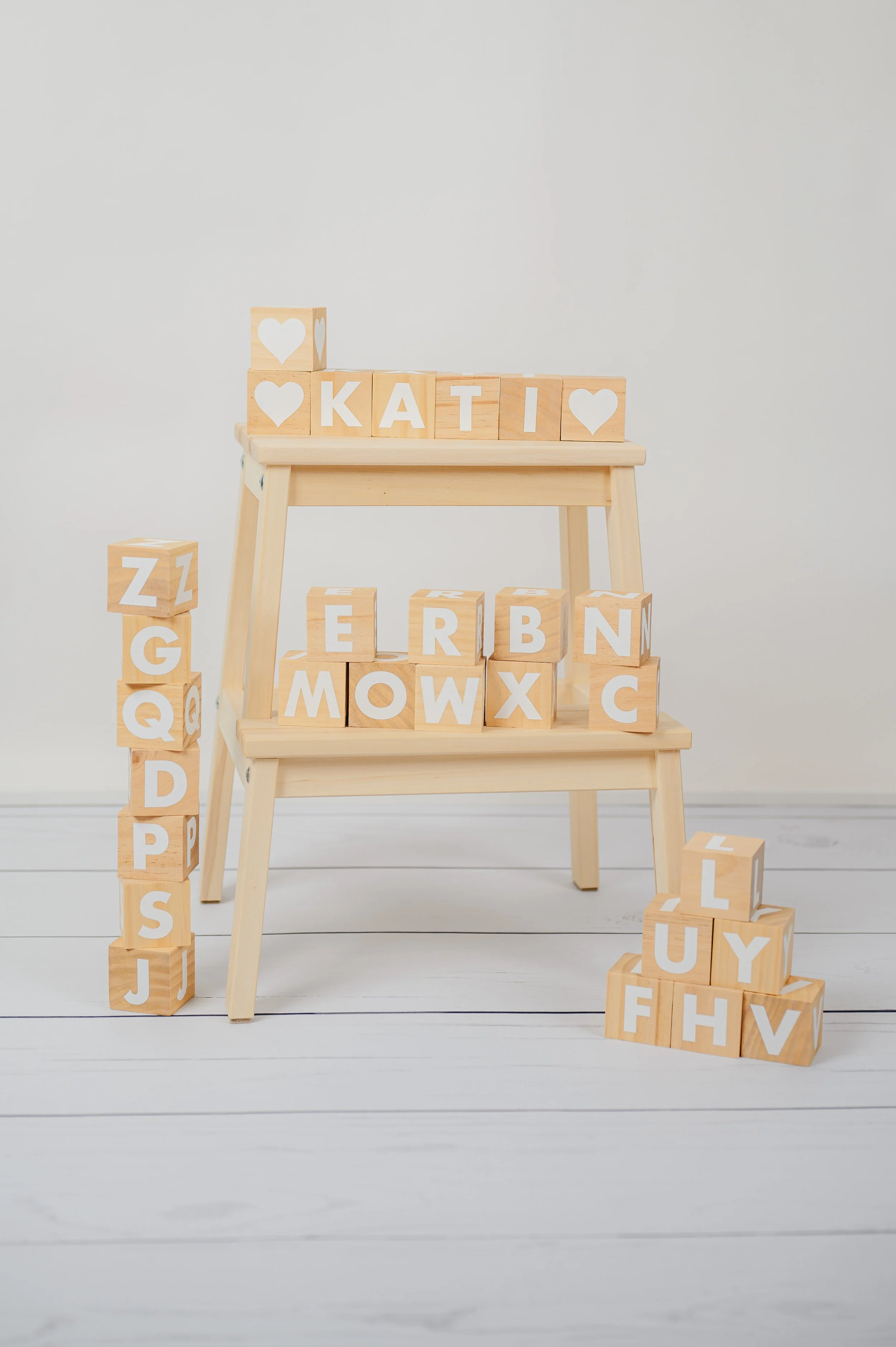 Natürliches Stillleben aus der Kita- und Schulfotografie mit Holzwürfeln: Holzbuchstaben zum Stapeln und Legen von Namen auf einem kleinen Hocker vor hellem Hintergrund. Ruhiges, modernes Fotoset für spielerische und kreative Kinderporträts.