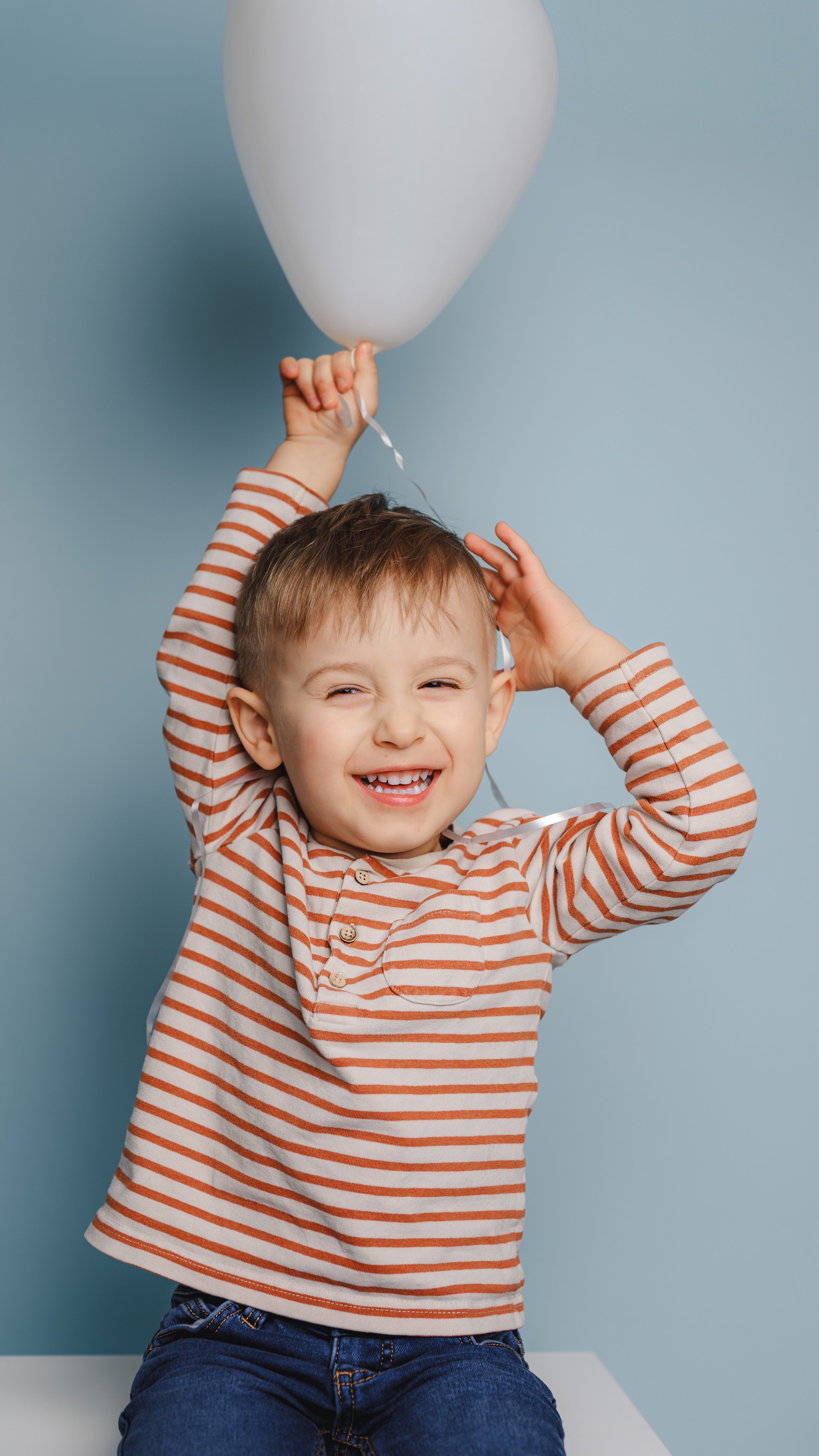 Fröhliches Kita-Foto eines Kindes mit weißem Luftballon vor blauem Hintergrund. Das spielerische Lachen und die natürliche Bewegung stehen für lebendige und kindgerechte Kita-Fotografie.