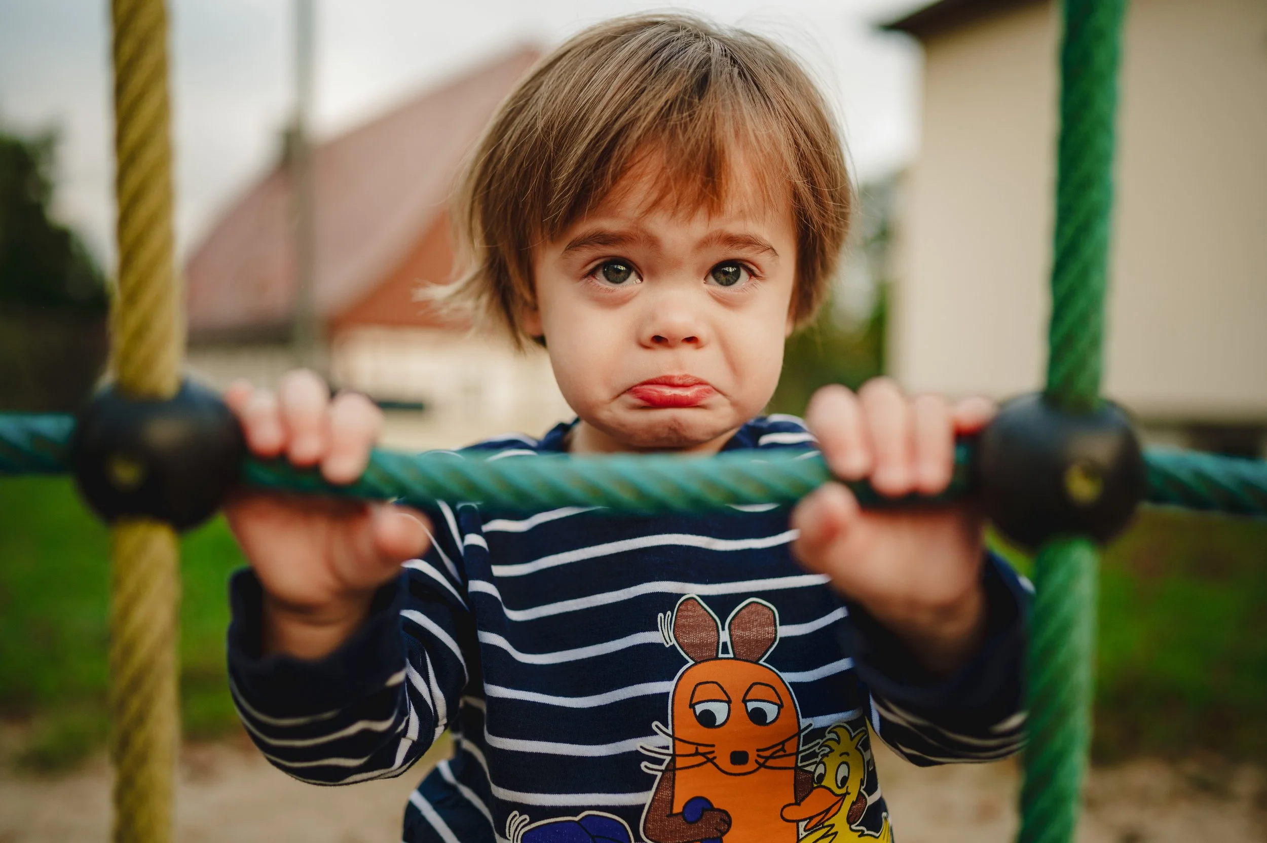 Natürliches Outdoor-Kita-Foto eines Jungen auf dem Spielplatz. Das Kind zeigt eine ehrliche, ungefilterte Mimik und wird bei weichem Tageslicht fotografiert. Ruhiger Hintergrund, echte Emotionen und eine entspannte Atmosphäre – moderne Kita-Fotografi