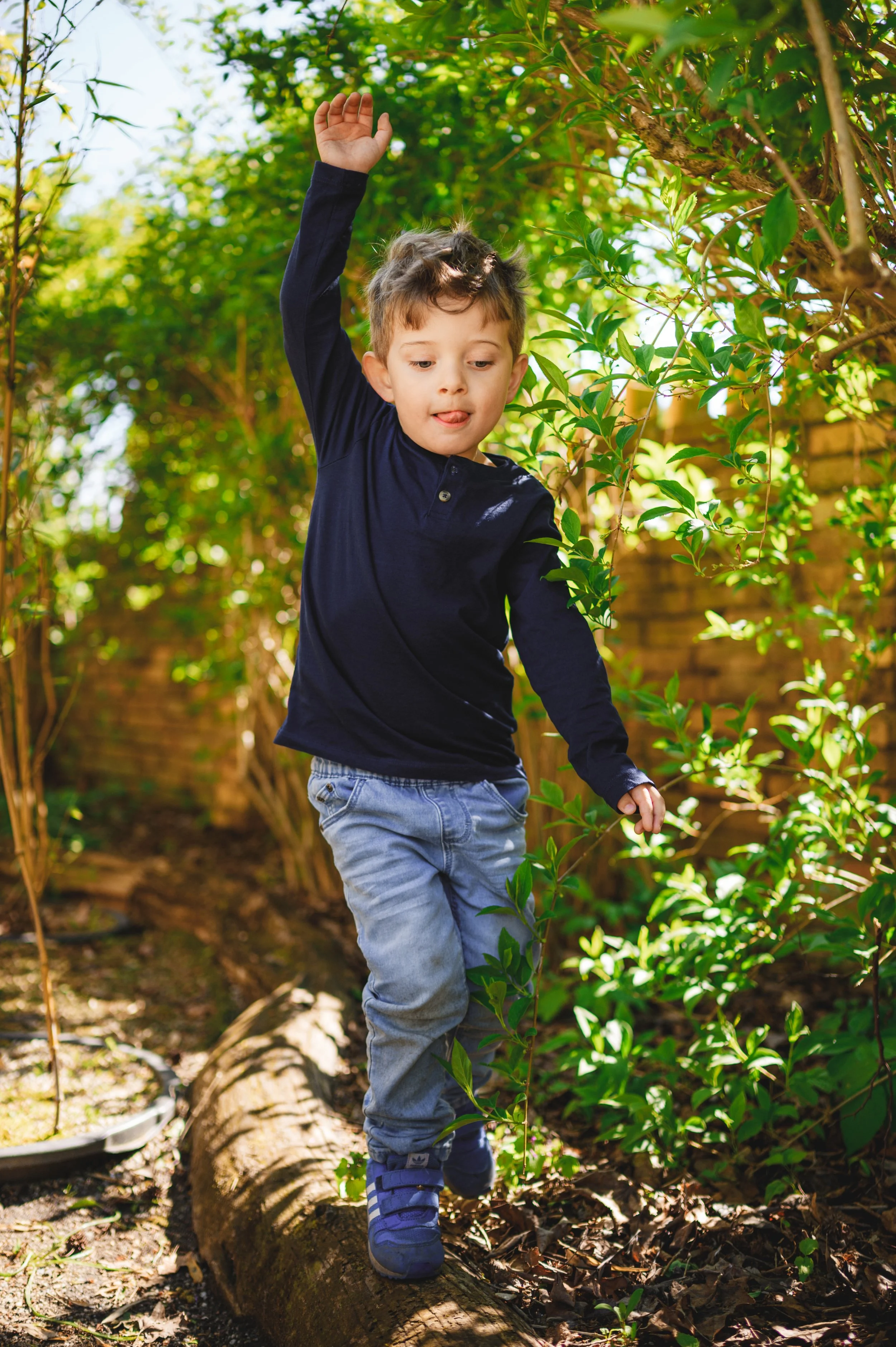 Ein Kita-Kind balanciert konzentriert über einen Baumstamm im Garten. Natürliche Outdoor-Kita-Fotografie mit Bewegung, echtem Moment und weichem Tageslicht.