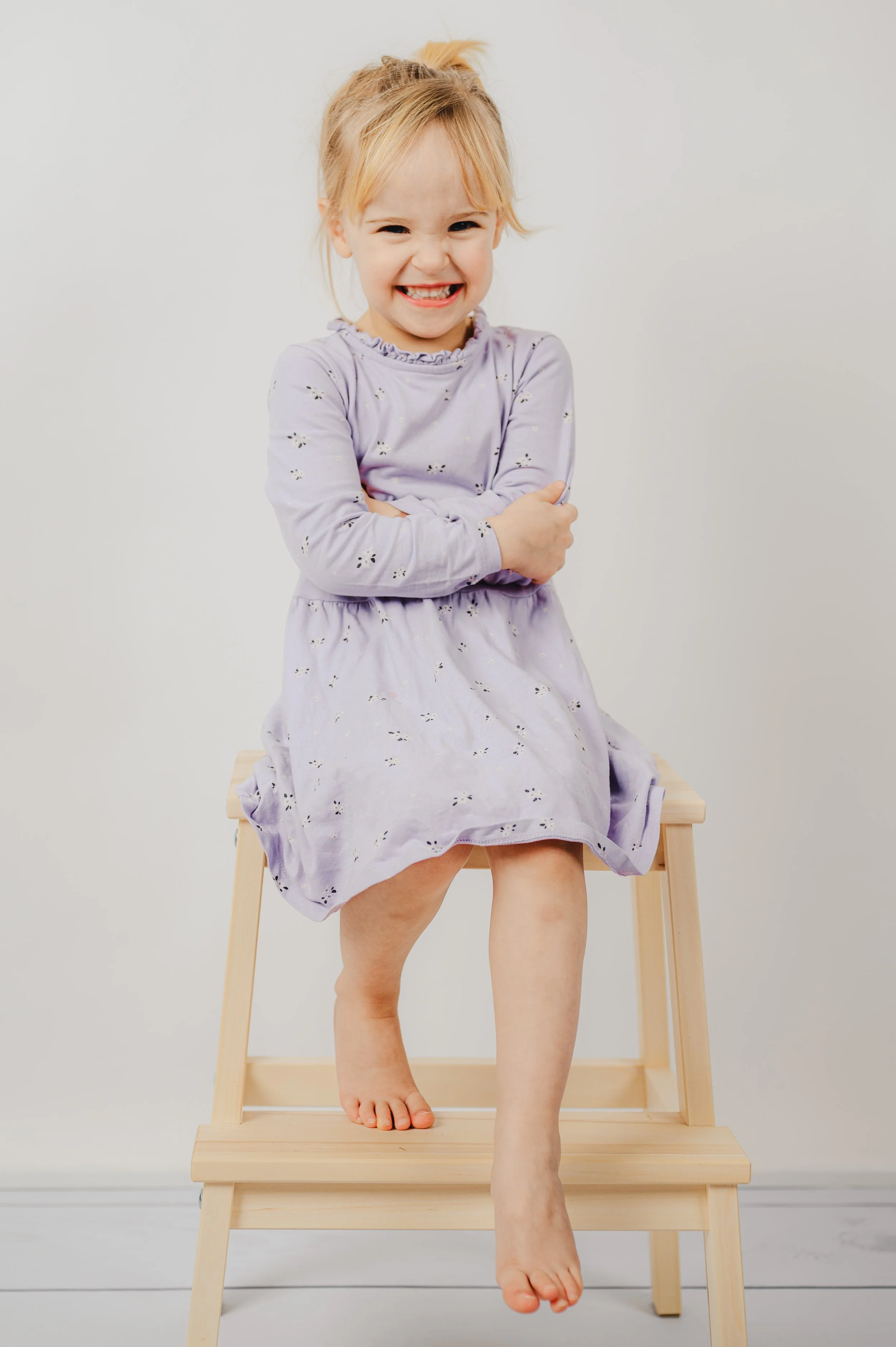 Natürliches Kita- und Schulfoto eines Kindes, das lachend auf einem Holz­hocker sitzt. Der helle, ruhige Hintergrund und die entspannte Haltung sorgen für eine moderne, zeitlose Bildwirkung. Echte Emotionen, natürliche Farben und eine spielerische At