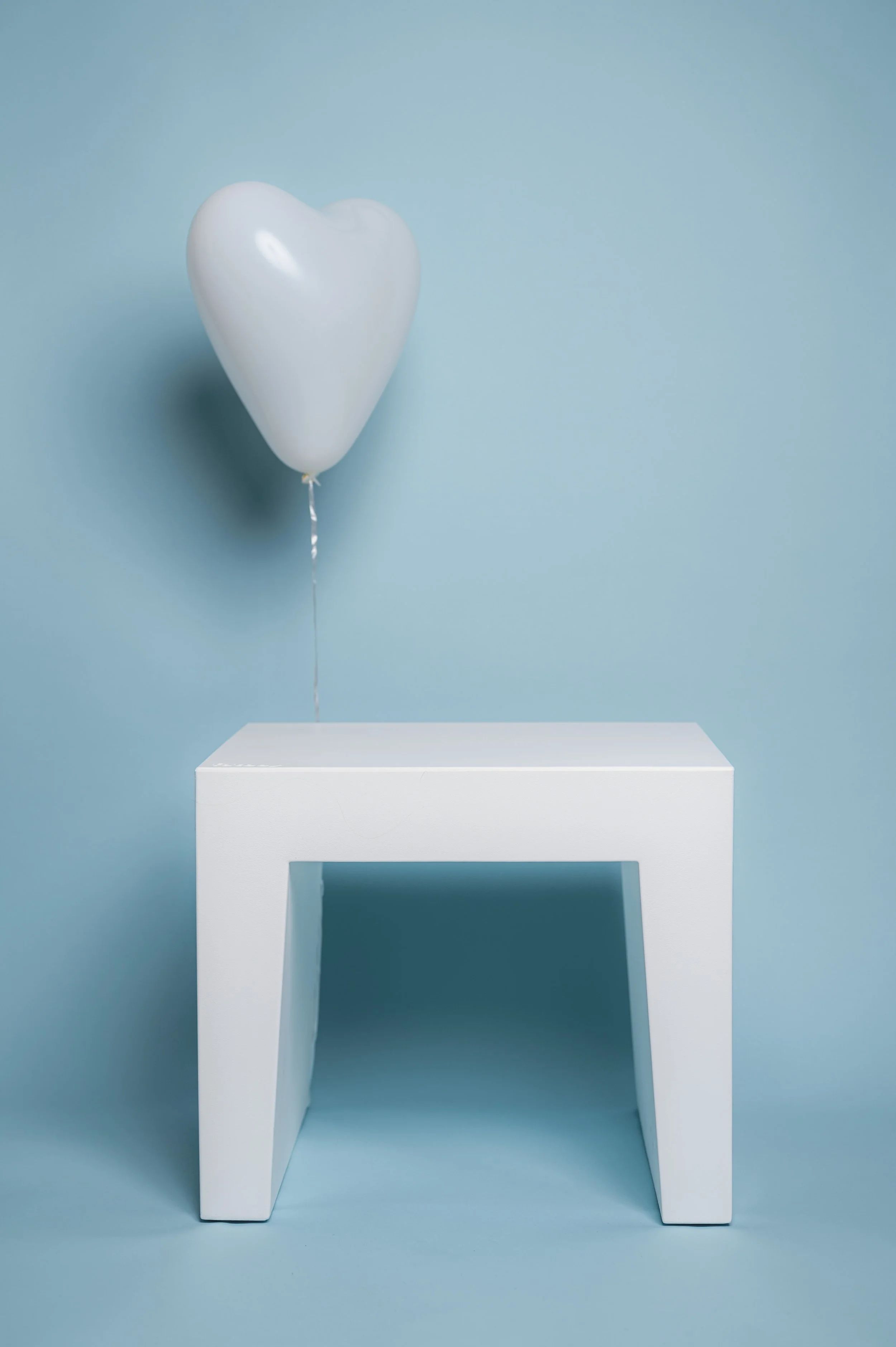 Minimalistisches Fotoset für die Kita-Fotografie: Ein weißer Luftballon schwebt über einem schlichten weißen Hocker vor blauem Hintergrund. Das luftige Set schafft eine ruhige, freundliche Atmosphäre und eignet sich ideal für natürliche und unbeschwe