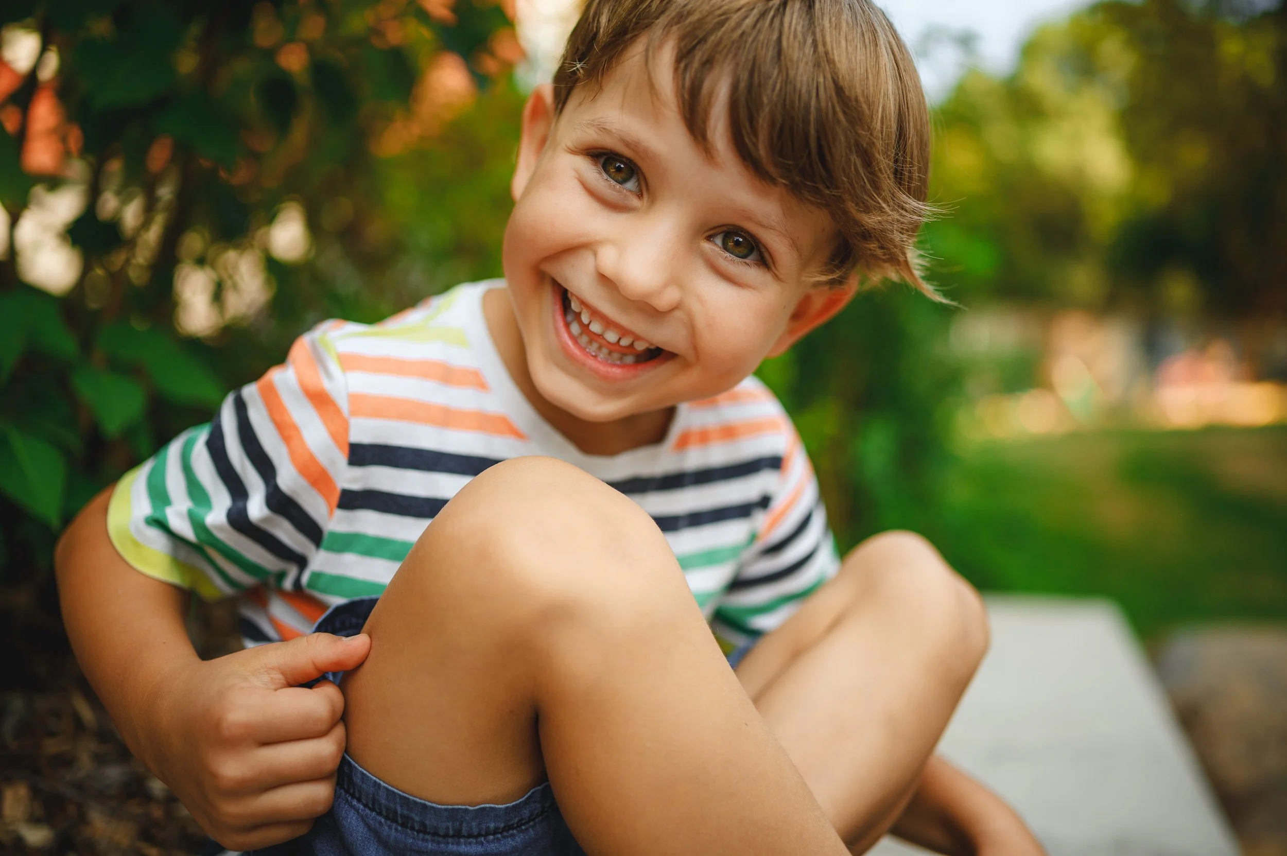 Ein fröhliches Kita-Kind sitzt entspannt im Grünen und lacht direkt in die Kamera. Authentische Outdoor-Kita-Fotografie mit natürlichem Licht und echter Lebensfreude.