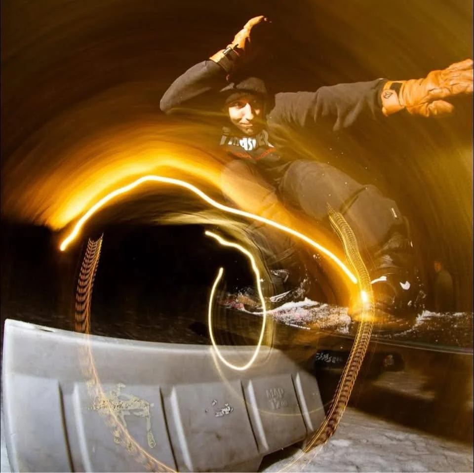 Person wearing a beanie hat and gloves, kneeling on the ground, with lights creating motion blur around them in a circular pattern at night.