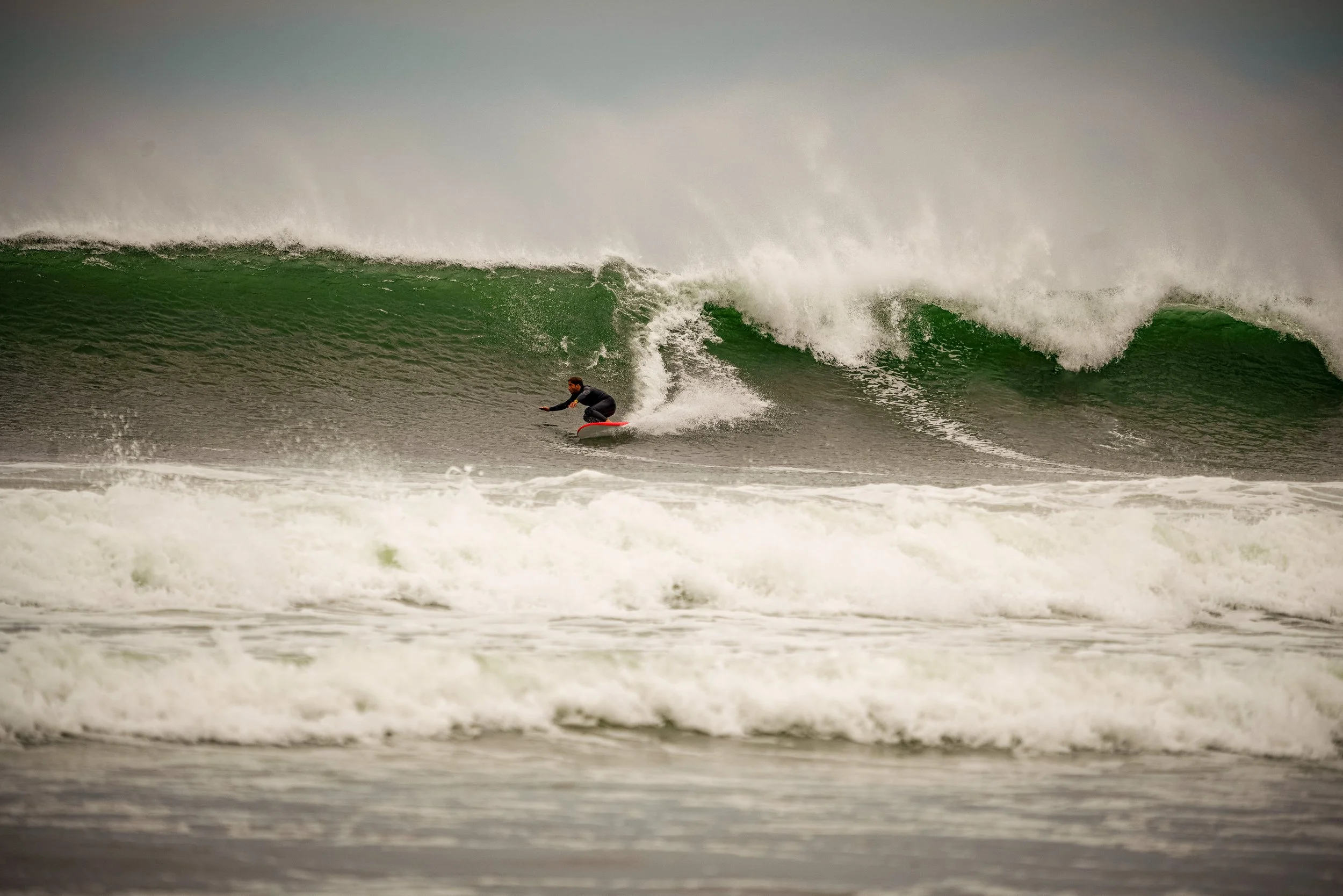 A person wearing a wetsuit surfing on a large green wave with white foam at the beach.