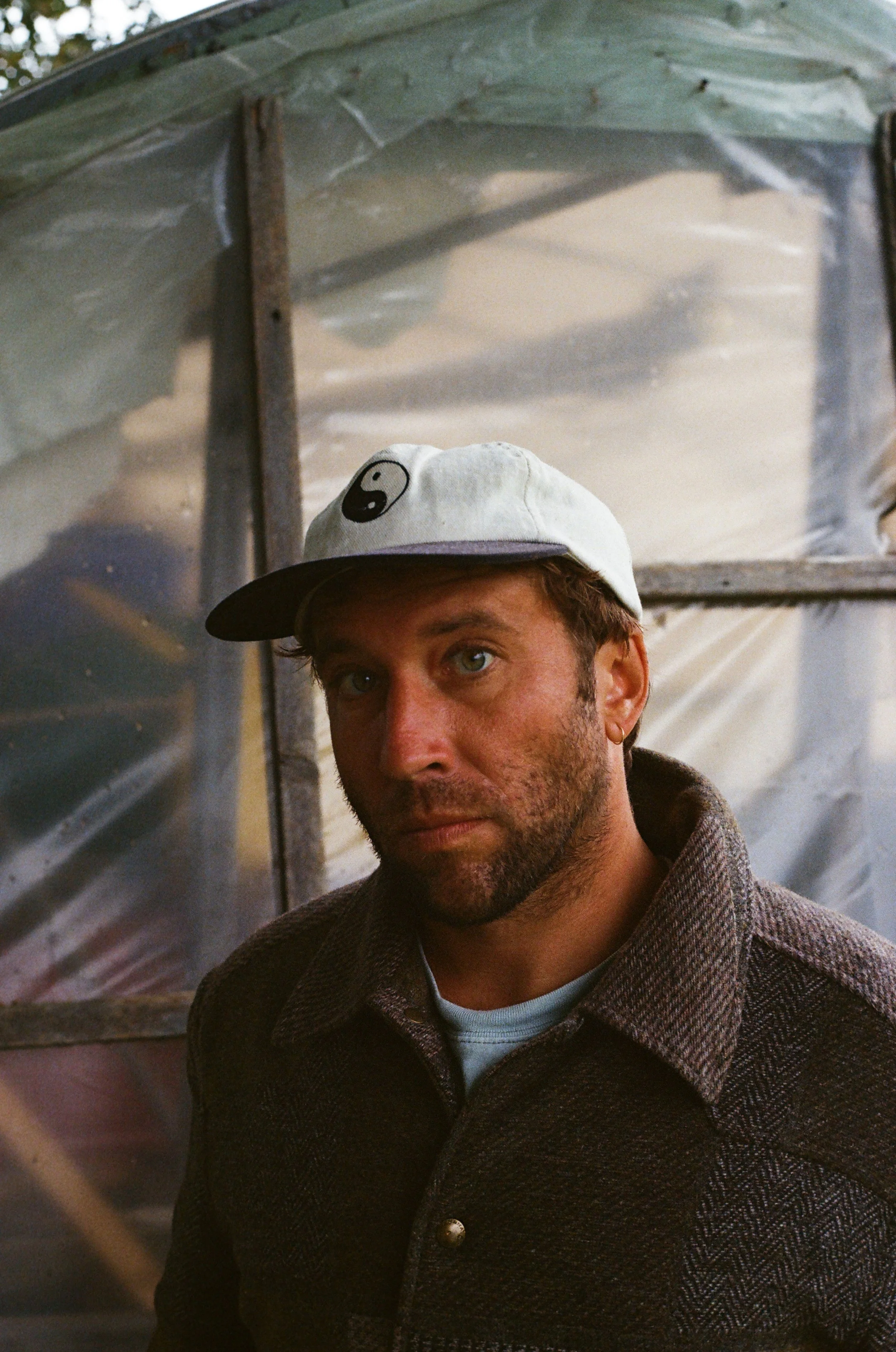 A man with light skin, green eyes, and brown hair wearing a white cap with a yin-yang symbol and a dark brown textured coat, standing in front of a reflective, metallic surface, possibly a trailer or van with a window.